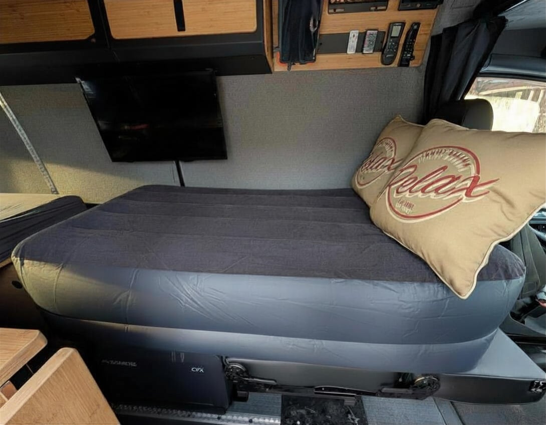 RV rental interior: Cozy air mattress bed with "Relax" pillows and a TV mounted in a comfortable camper van setup.. Winnebago Adventure Wagon 2023