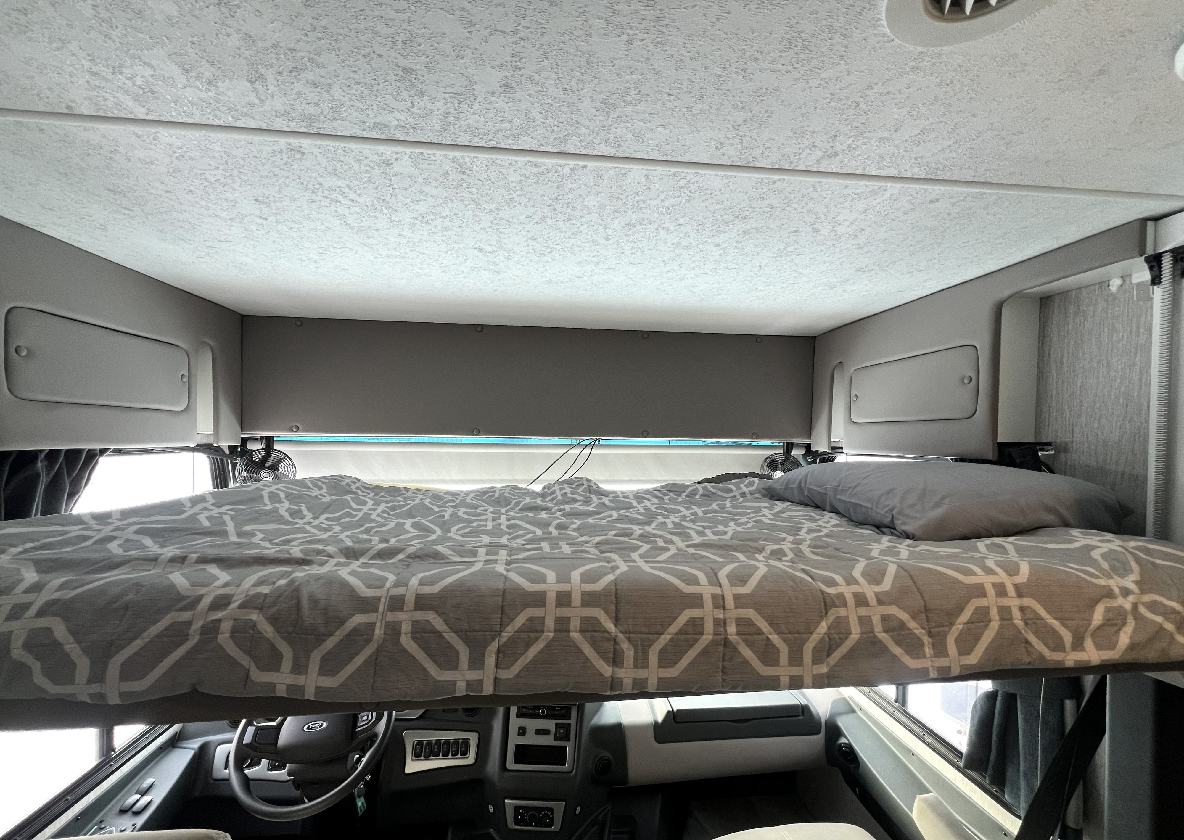 Cozy overhead bunk in an RV rental, featuring a grey patterned comforter, pillow, and small fans above the driver's cabin.. Coachmen Encore 2021