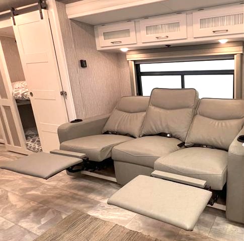 Spacious RV rental interior with comfy recliner seating, overhead storage, and barn door leading to a bunk area. Ideal for travel.. Coachmen Encore 2021