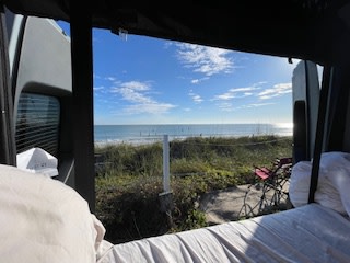 Wake up to this amazing ocean view from an RV rental! Inside a camper, you see blue sky, waves, beach grass, and a red chair by the sunny sea.. Winnebago Revel 2026
