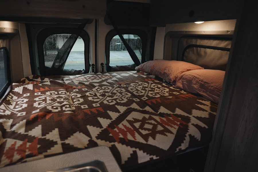 RV rental interior: A cozy bed with a patterned blanket, pink pillows, and windows, perfect for mobile sleeping.. Winnebago Winnebago 2025