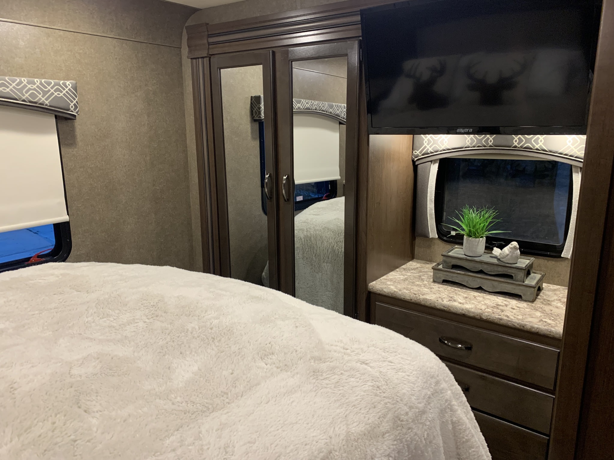Cozy RV rental bedroom: soft white comforter, mirrored closet, TV, and decorative items on a counter.. Thor Chateau 2020