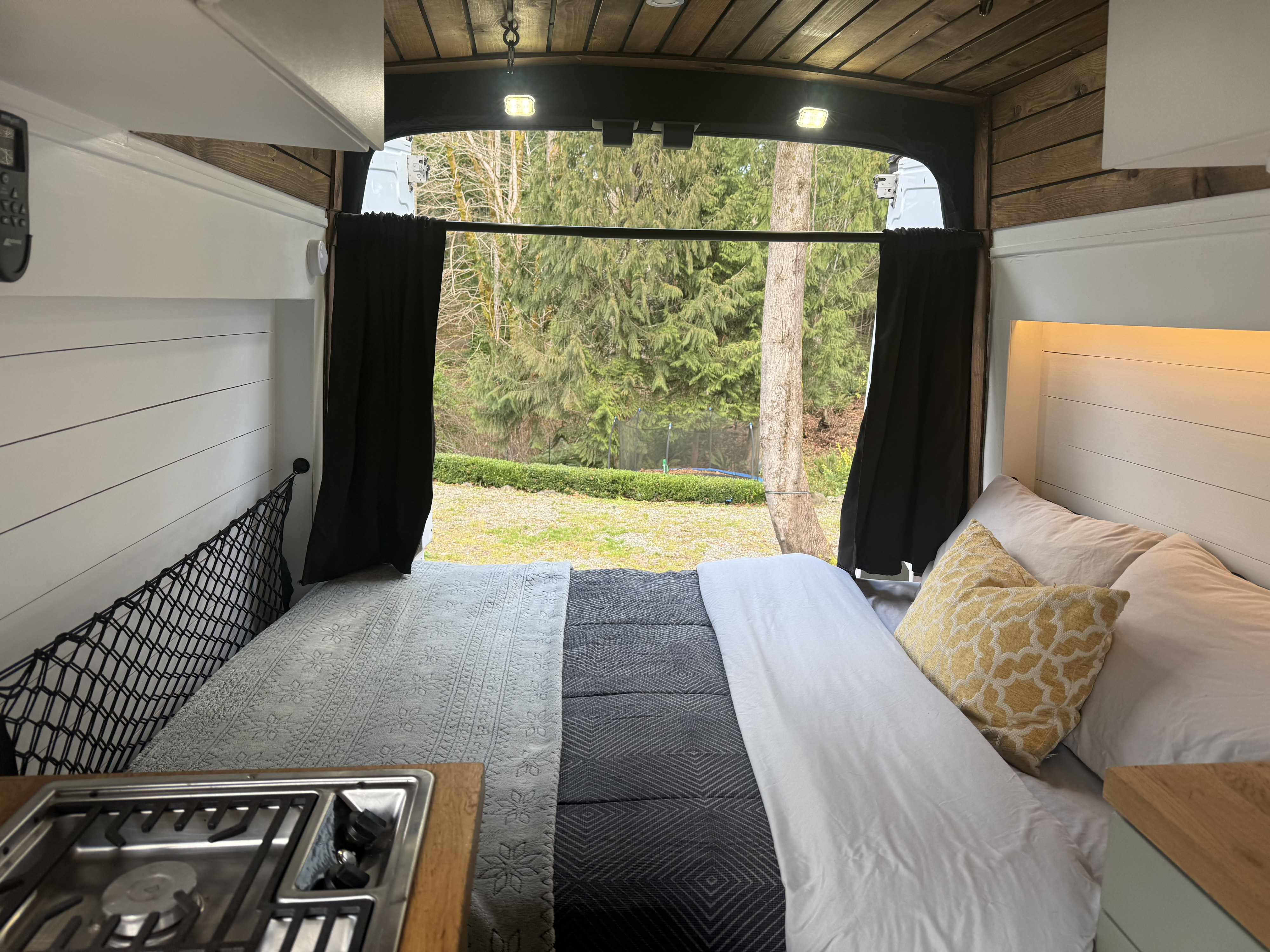 RV rental interior: cozy bed, compact kitchen stove, and serene forest view from open rear doors of this camper van.. Ford Transit 2015
