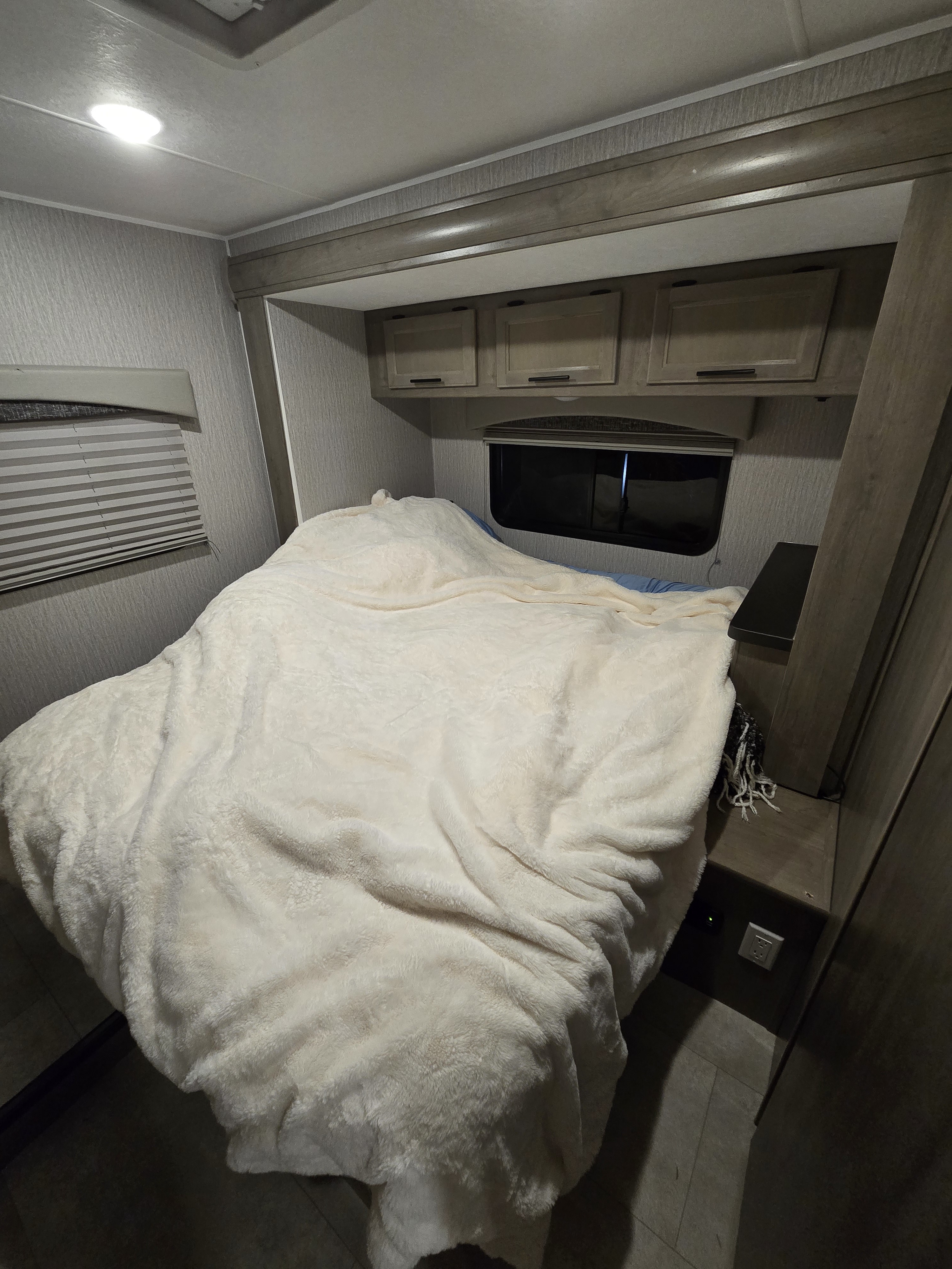 An RV rental's bedroom, featuring a large bed with a cream fluffy blanket, overhead cabinets, and a window.. Ford Coachmen Freelander Ford Coachmen Freelander 2023