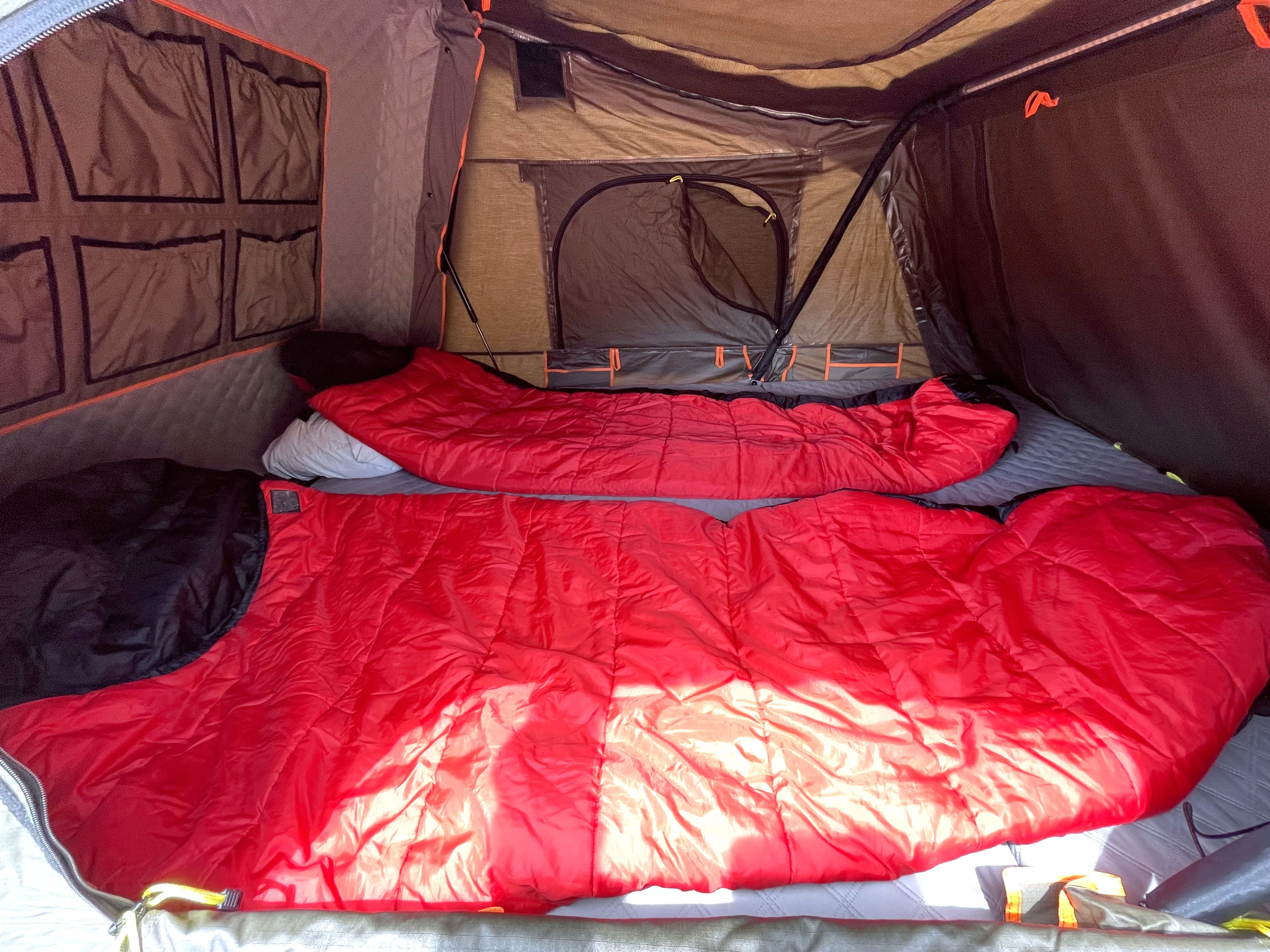 Inside a spacious RV rental tent: two red sleeping bags and pillows are laid out on a mattress, with storage pockets lining the wall.. Jeep Wrangler Unlimited 2020