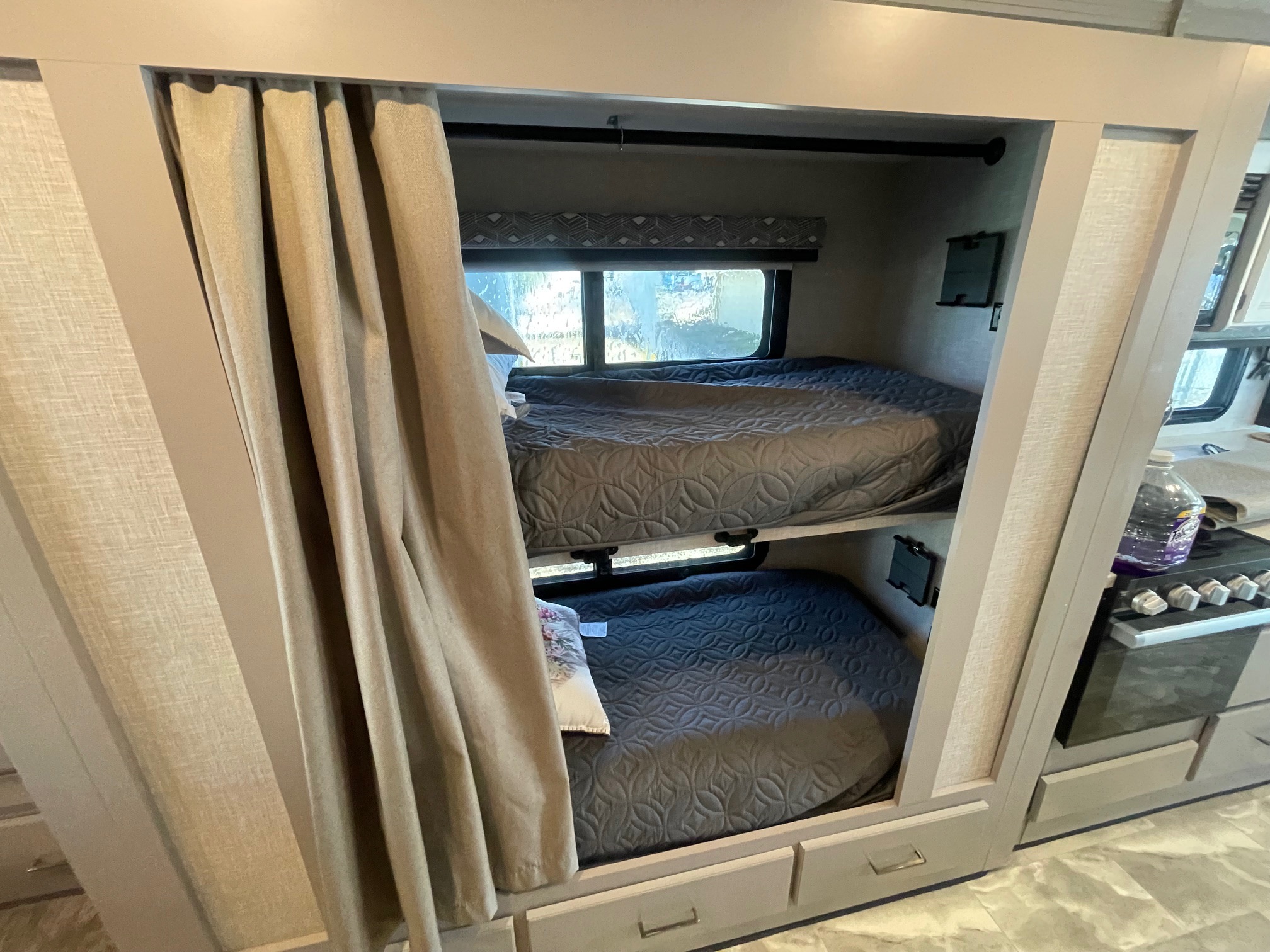 View of an RV rental's stacked bunk beds, featuring gray quilted bedding, windows, a privacy curtain, and storage drawers.. Thor Windsport 2024