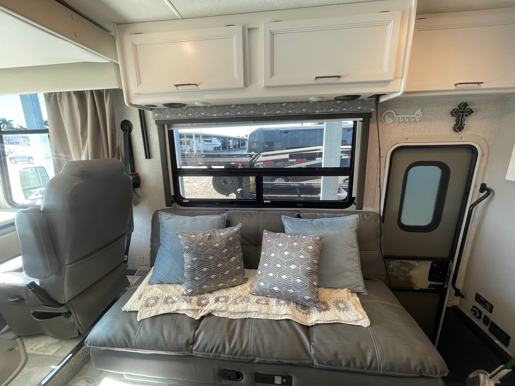 Cozy rv rental interior features a gray sofa with patterned pillows, overhead cabinets, large window, and driver's seat for travel.. Thor Windsport 2024