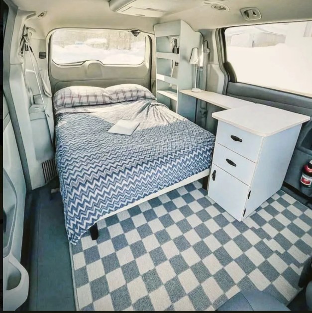 This RV rental's cozy camper van interior features a neatly made bed, a functional desk, and storage, all on a charming checkered rug.. Dodge B Van 2017