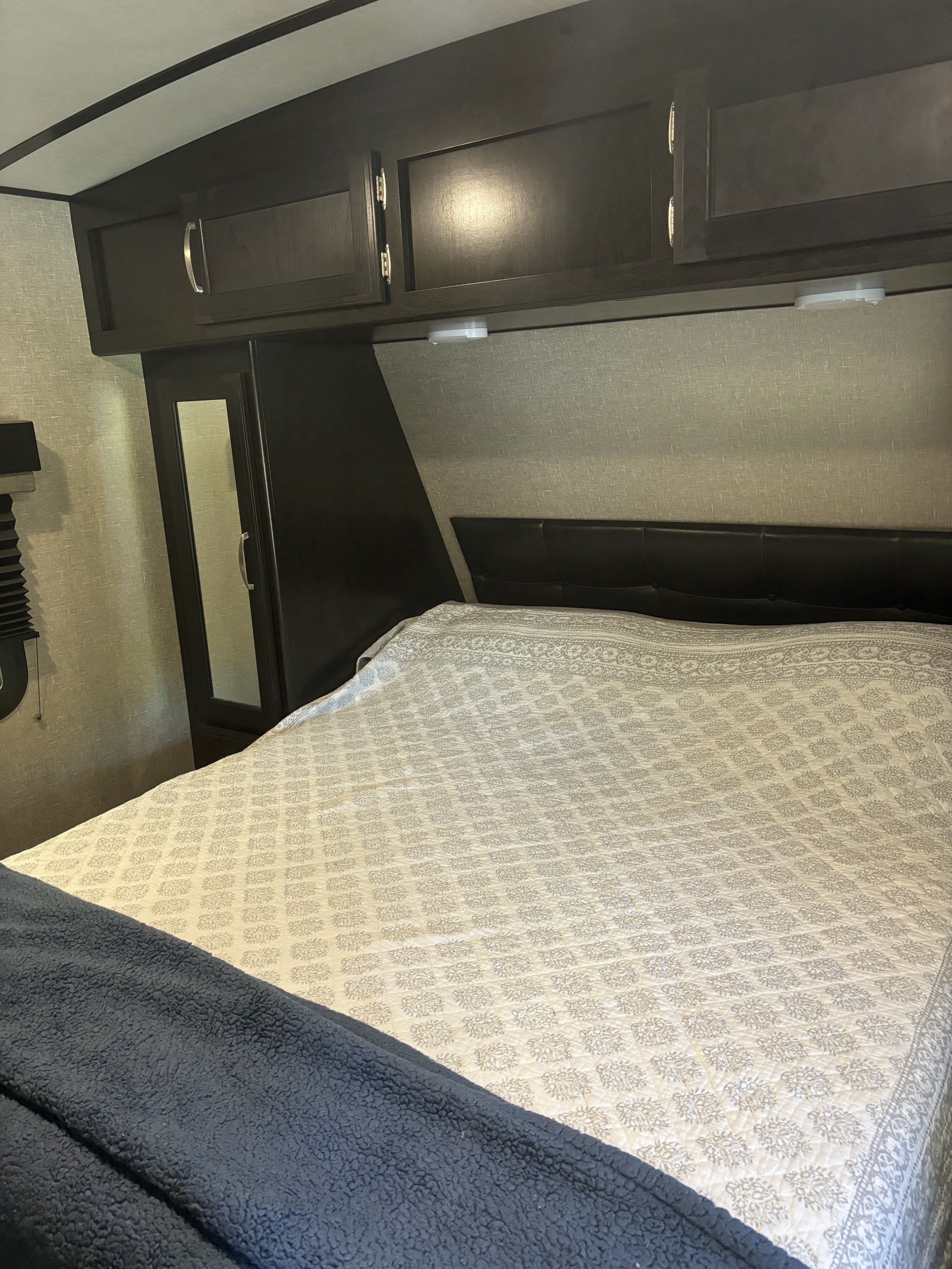 Inside an inviting rv rental bedroom, featuring a patterned bed, dark headboard, overhead storage, and mirrored wardrobe.. Grand Design Imagine 2019