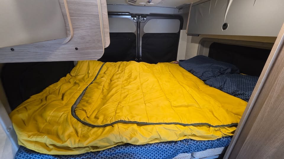Cozy bed in an RV rental, featuring a bright yellow sleeping bag, blue pillows, and overhead storage for comfortable travels.. Winnebago Solis 2023