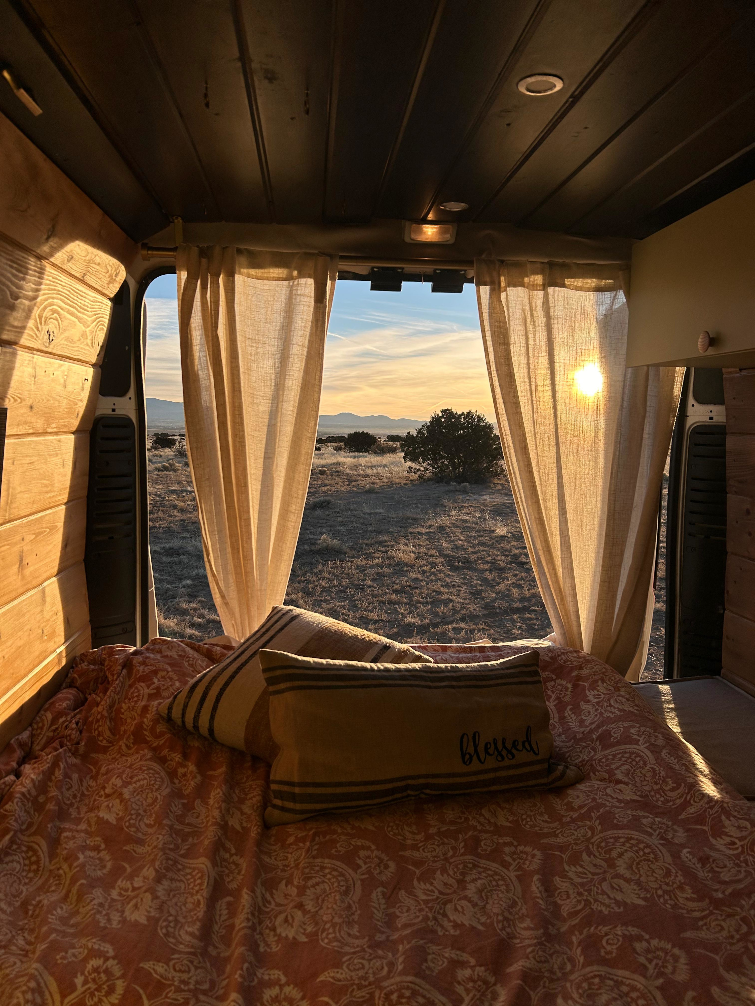 Cozy RV rental interior offers a stunning sunset view over a desert landscape through open back doors, framed by curtains and golden light.. RAM Promaster 2019