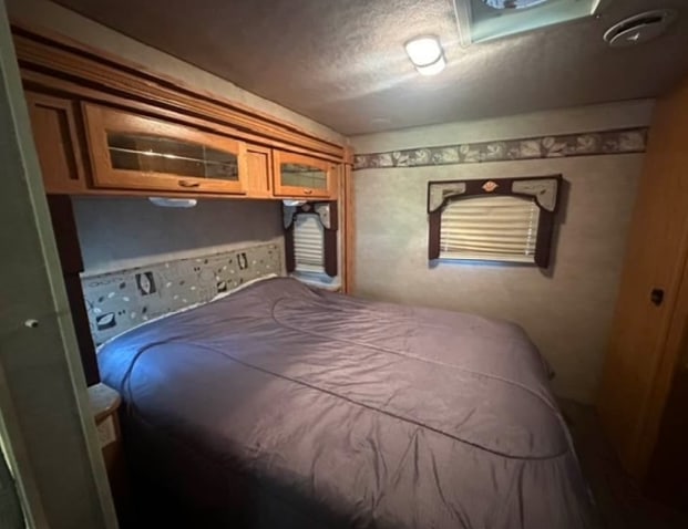 Inside an RV rental: a compact bedroom featuring a double bed with purple bedding, overhead storage, and a window.. Jayco Greyhawk 2006