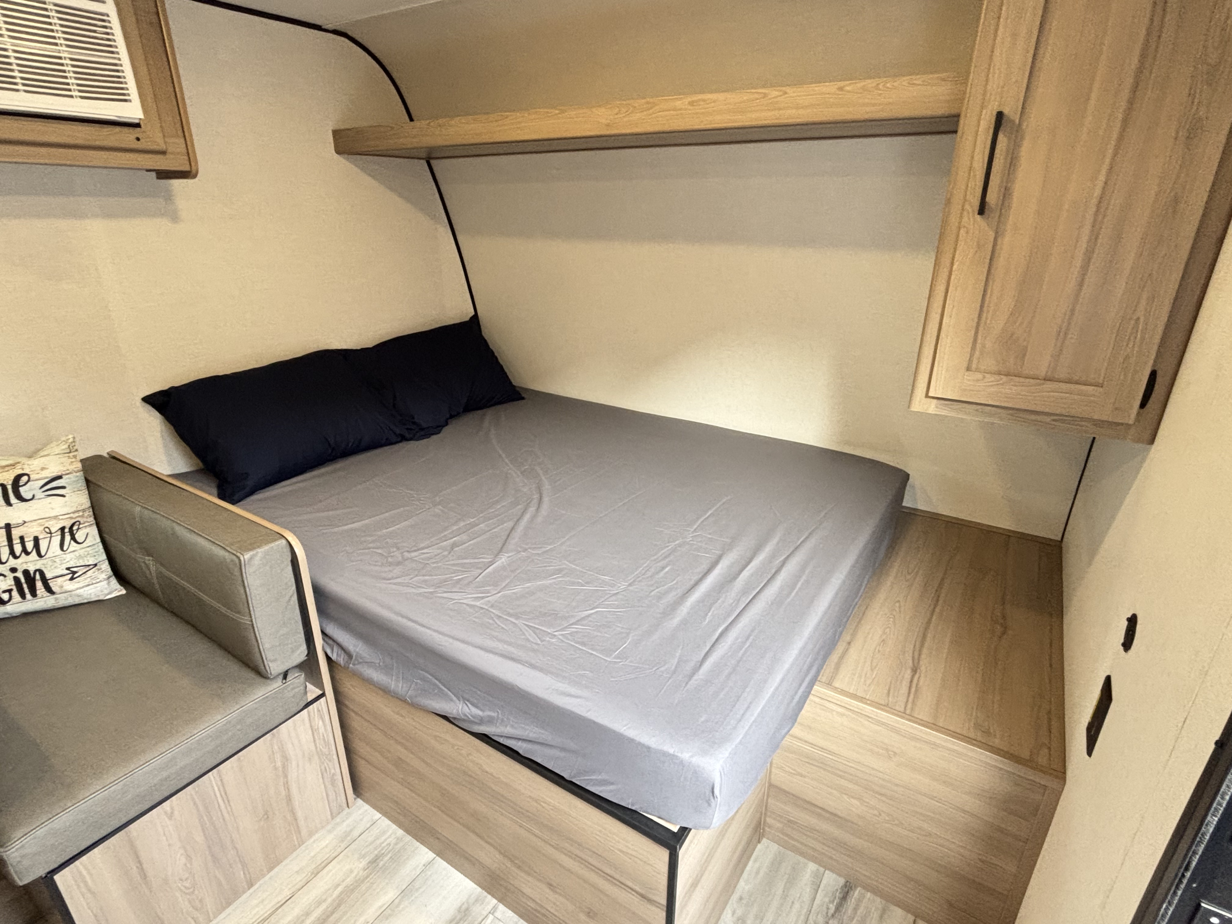 Cozy RV rental interior with a compact bed, gray sheets, dark pillows, sofa seating, overhead shelf, and AC unit.. Keystone Coleman 2026