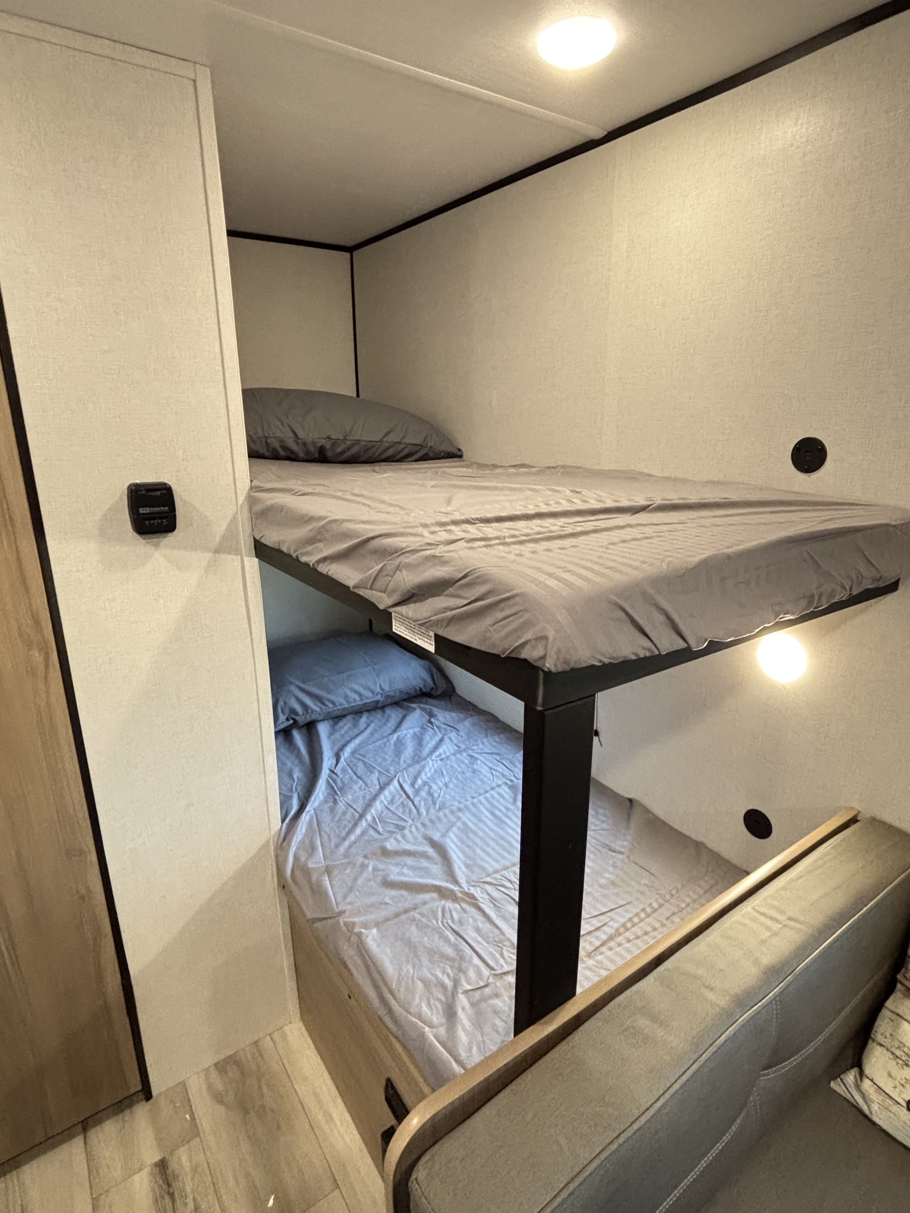 Inside an RV rental: two bunk beds with grey sheets, an overhead light, and a small reading lamp below the top bunk.. Keystone Coleman 2026