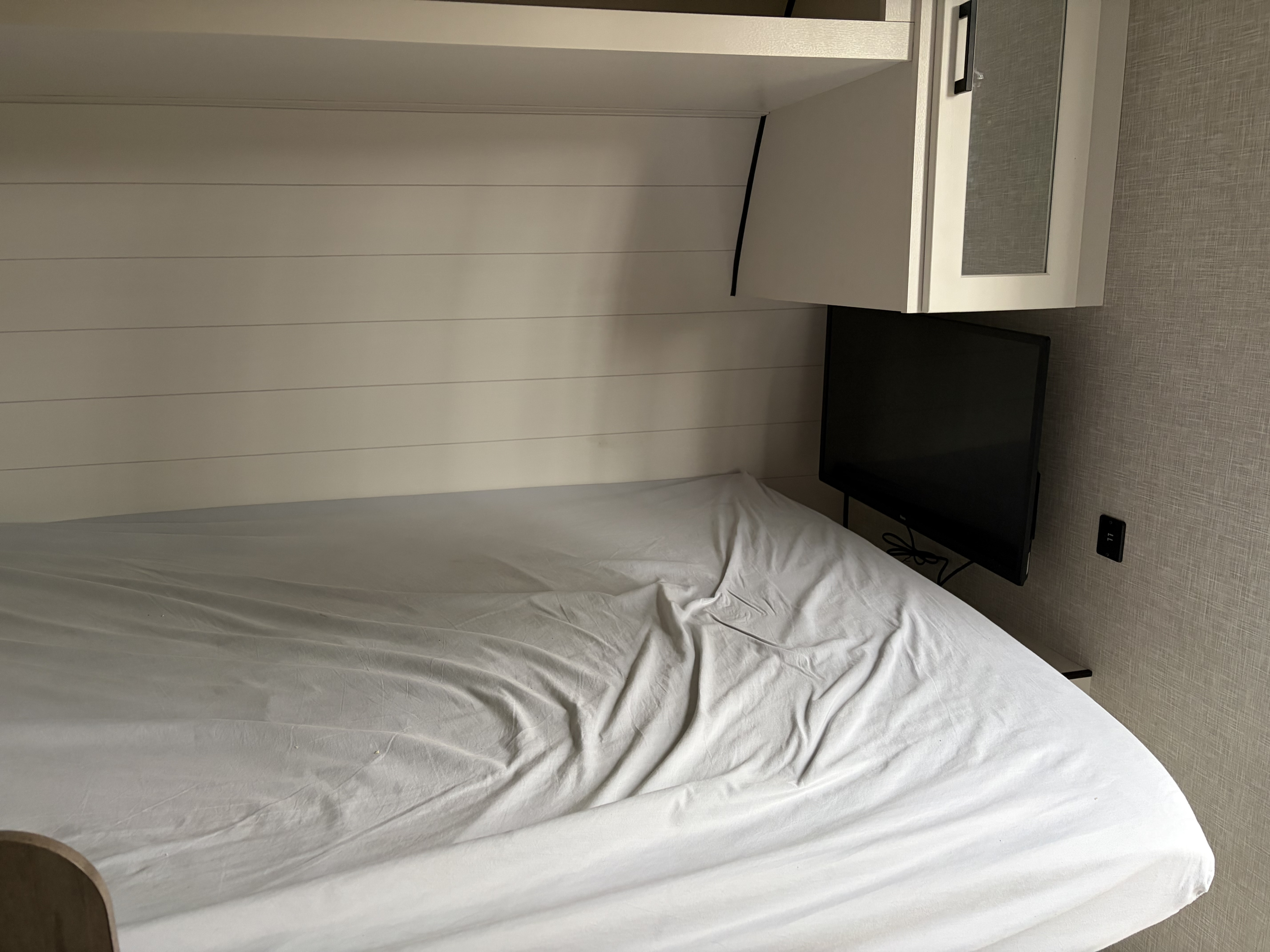 Cozy RV rental bedroom featuring a bed with white sheets, shiplap walls, wall-mounted TV, and overhead cabinet storage.. Jayco Jay Flight SLX 2025