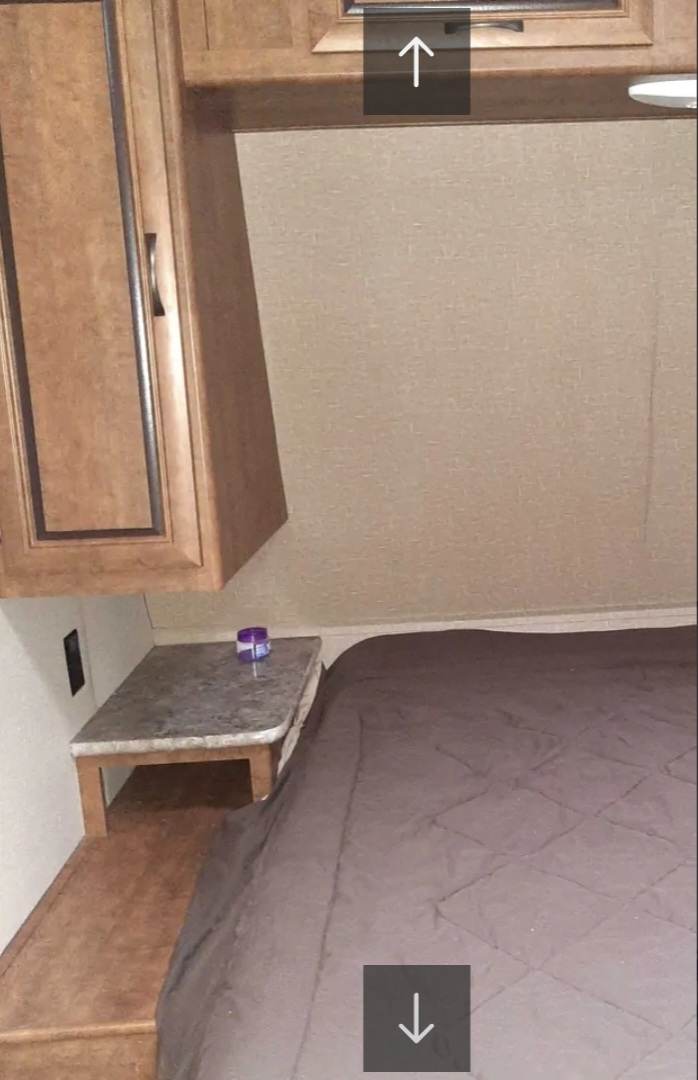 RV rental bedroom with a cozy bed, small nightstand holding a purple container, and wooden overhead cabinets.. Crossroads RV Zinger 2017