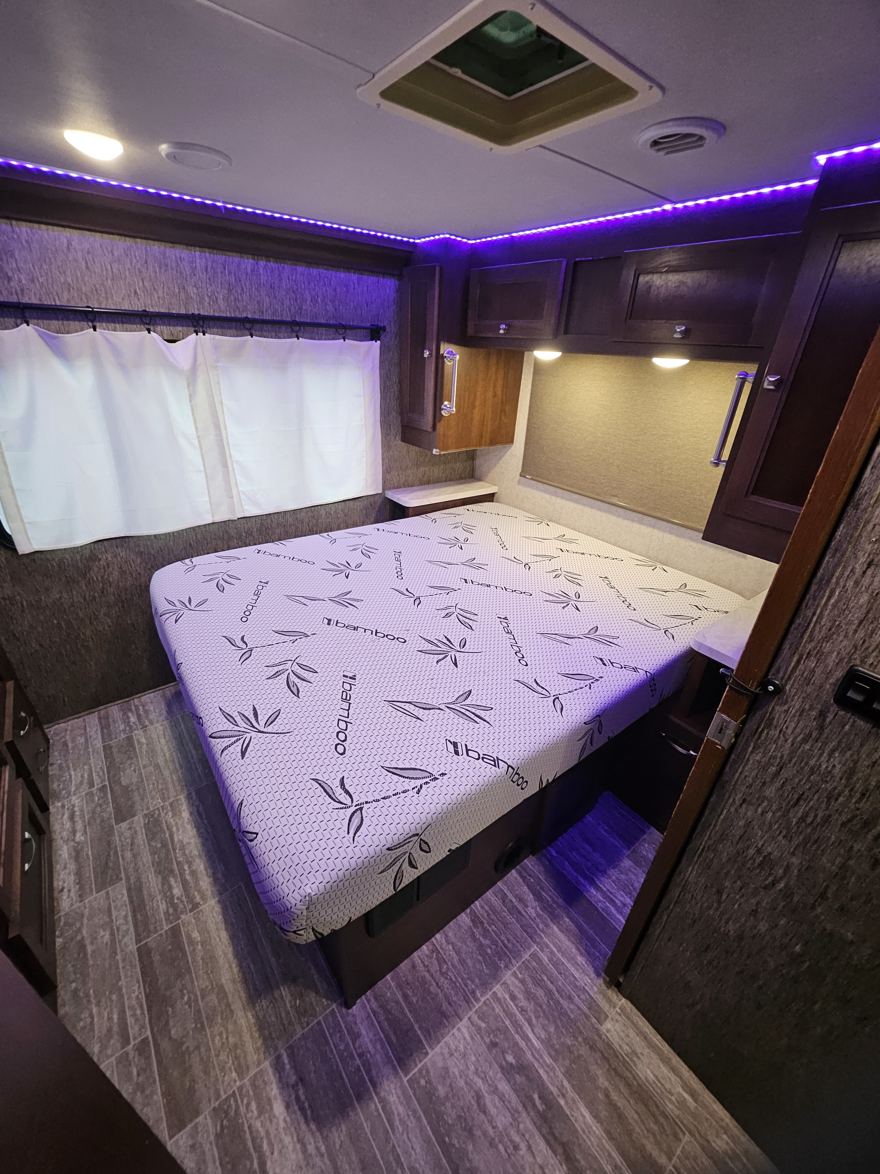 RV rental bedroom featuring a bamboo mattress, dark wood cabinets, purple accent lighting, a window with curtains, and wood-look flooring.. Forest River Forester 2018