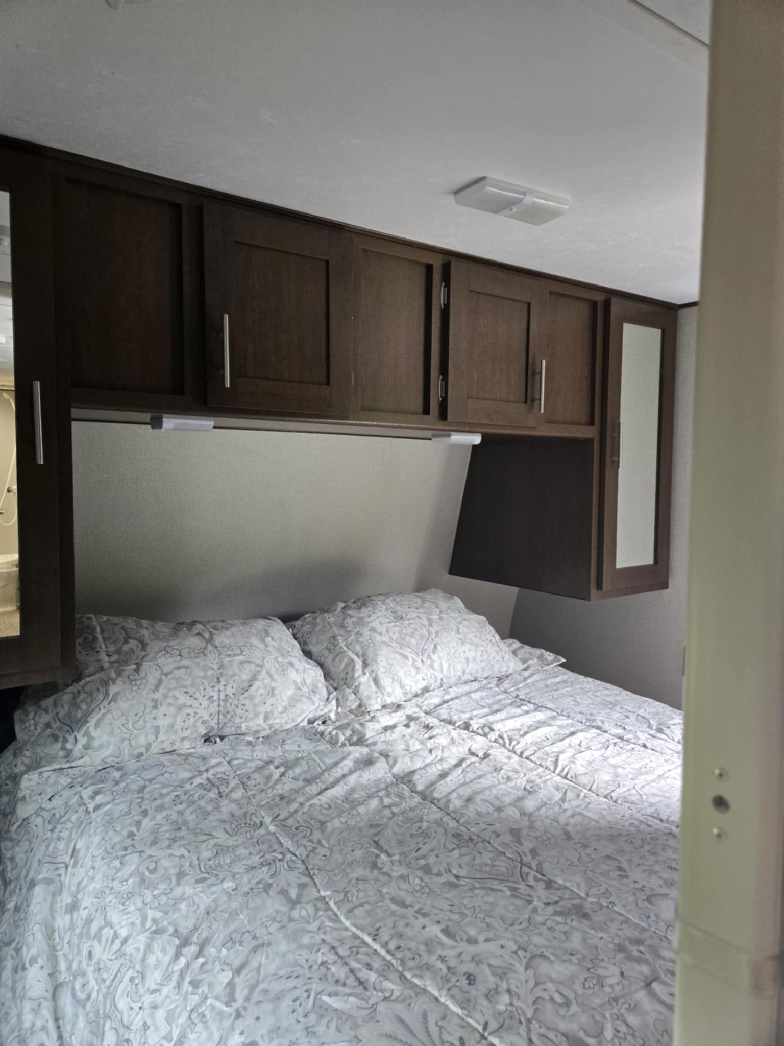 Bright RV rental bedroom features a bed with white and gray patterned bedding, dark wood overhead cabinets, and recessed lighting.. Avenger ATI 2019