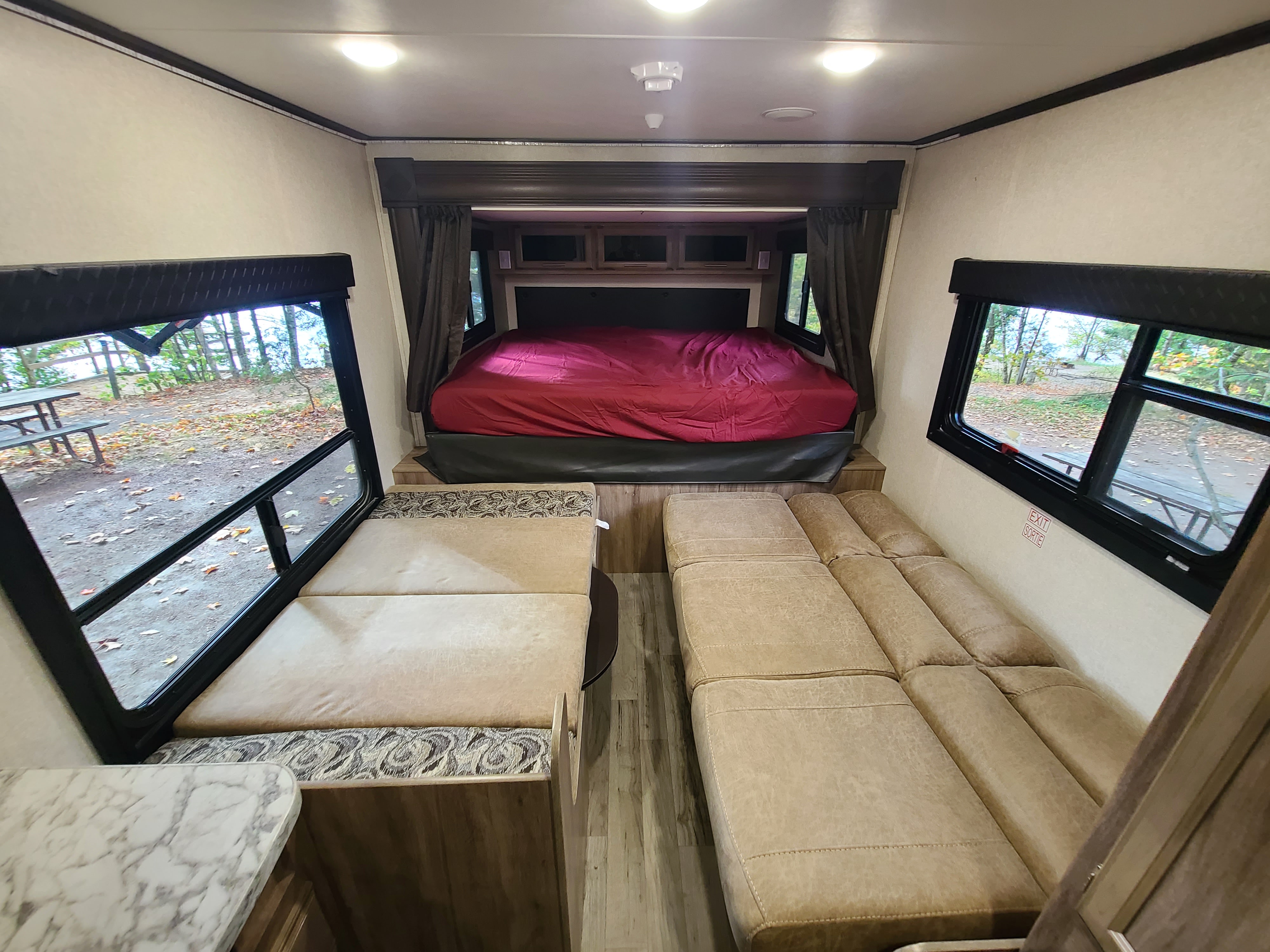 Cozy RV rental interior showing a bed with red cover, two cushioned seating areas, and windows revealing an outdoor campsite.. Jayco Jay Feather 2018