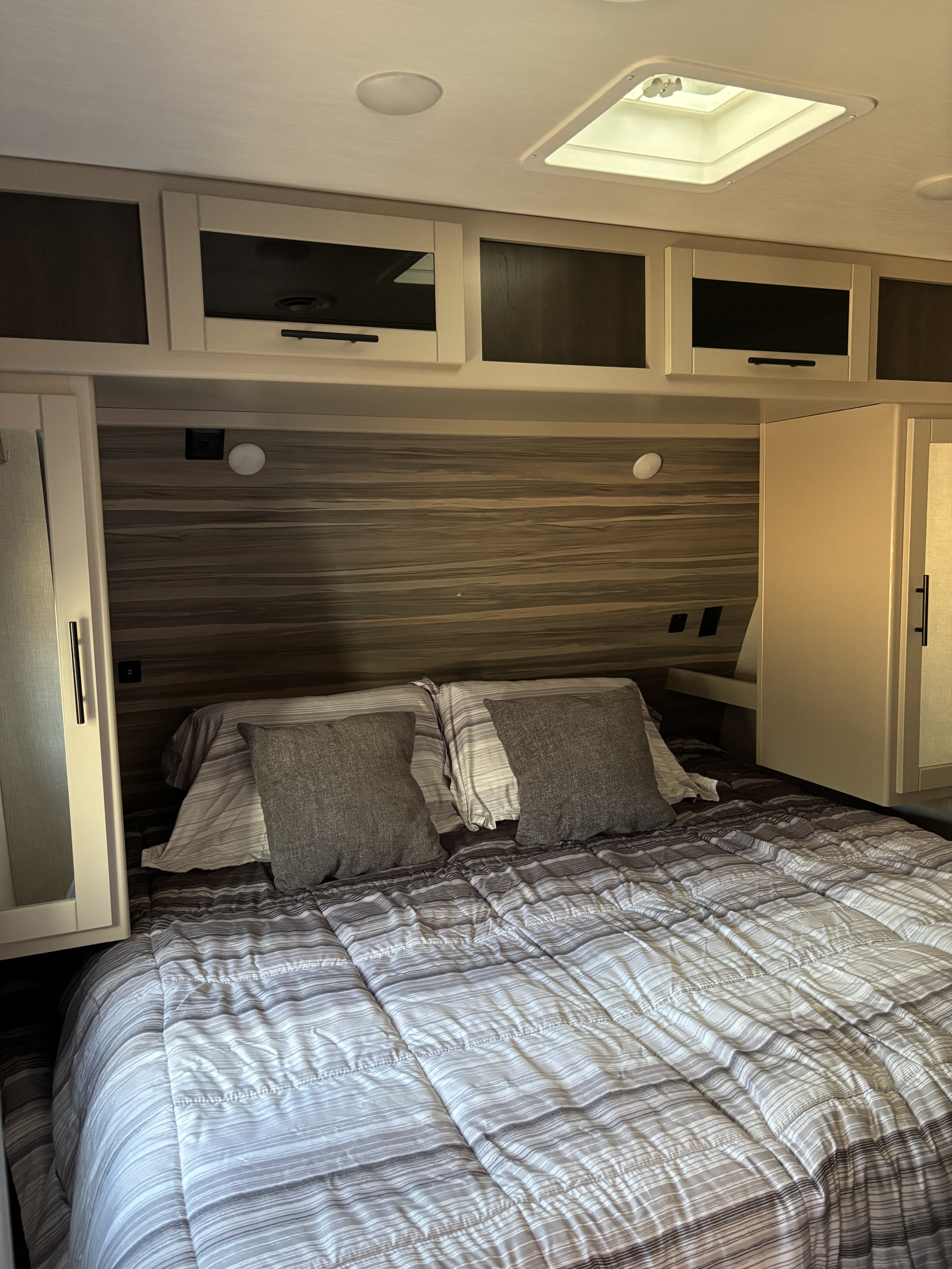 Cozy RV rental bedroom featuring a bed with striped bedding, overhead storage cabinets, a wood-paneled headboard, and a skylight.. Crossroads RV Cruiser Aire 2022