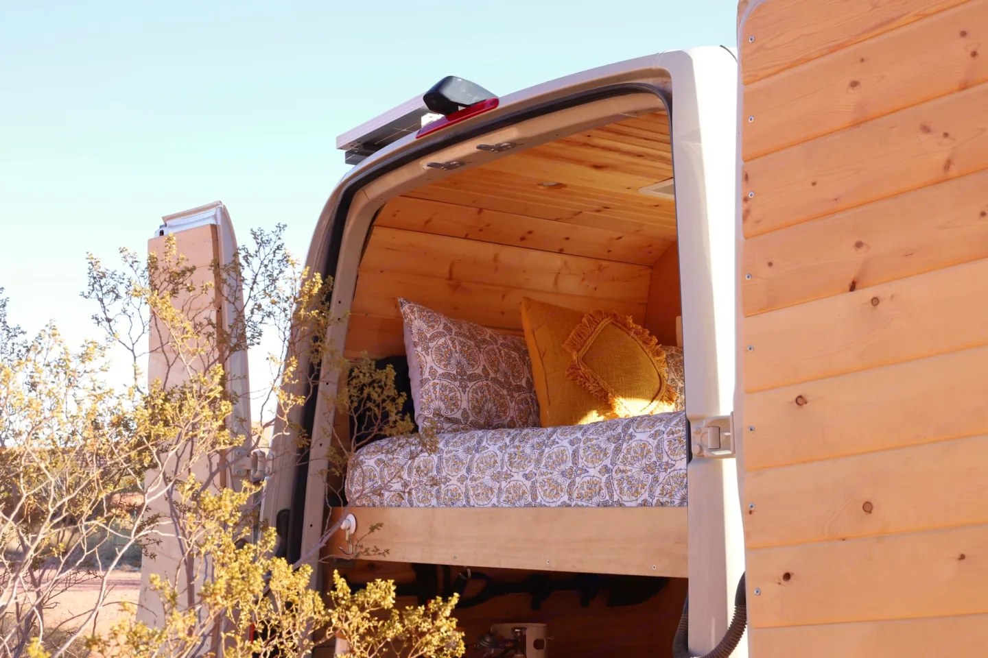 An RV rental's cozy custom wood-paneled interior, featuring a bed with patterned pillows, viewed through its open back doors. Ideal for van life.. Mercedes sprinter 2019