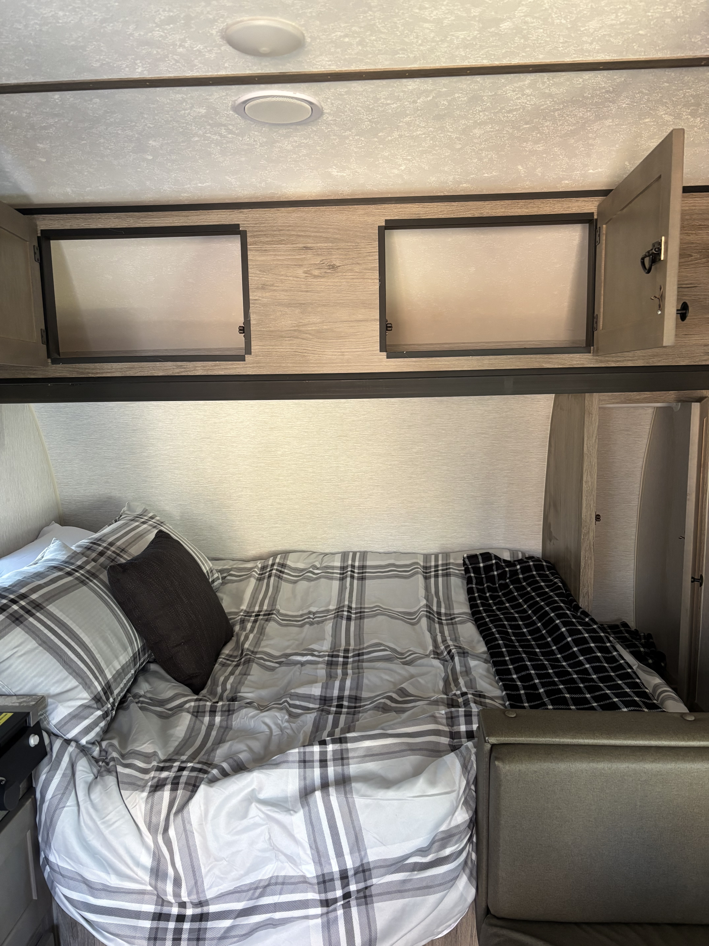 An RV rental bed featuring gray plaid bedding, a dark pillow, and open wooden overhead cabinets with a textured ceiling.. Sunset Park RV Sun Lite 2023