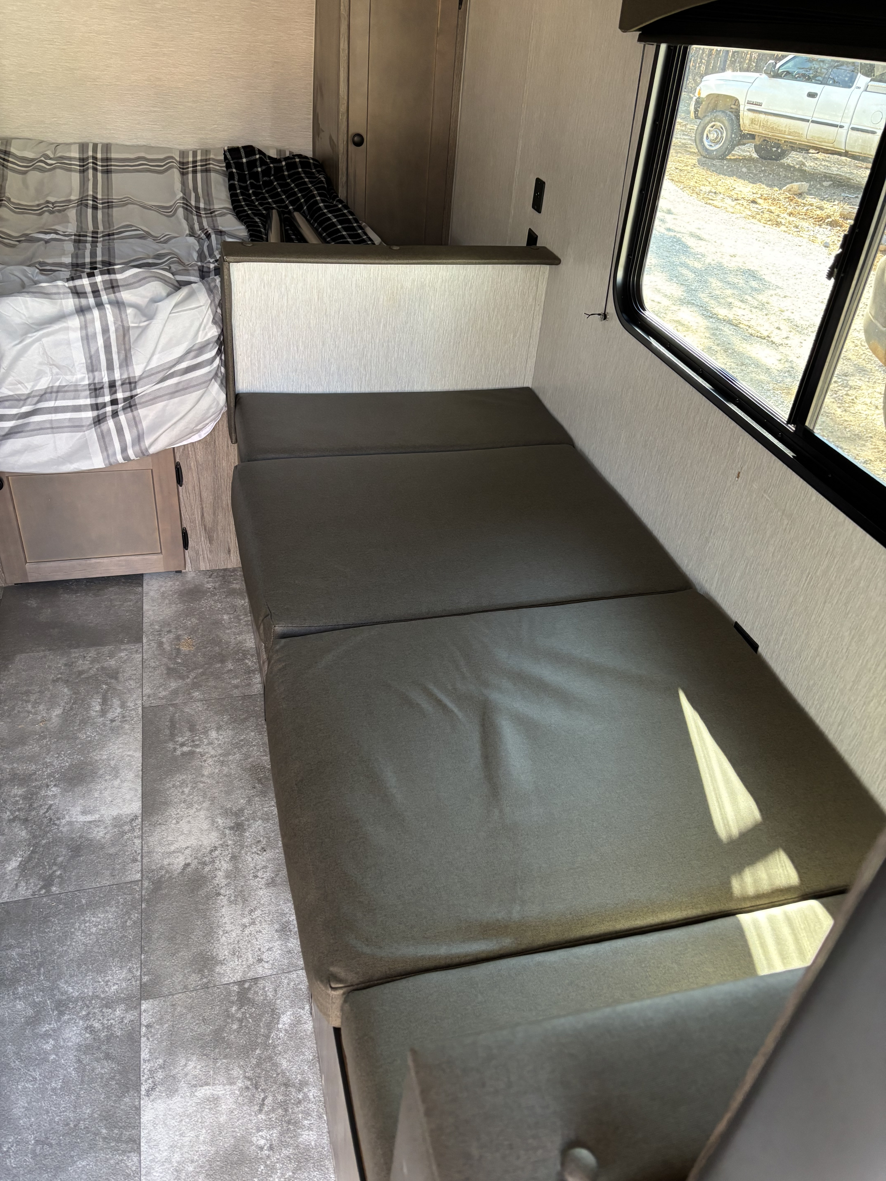 RV rental interior with a plaid bed, dark grey bench seating, and a window revealing a truck outside.. Sunset Park RV Sun Lite 2023