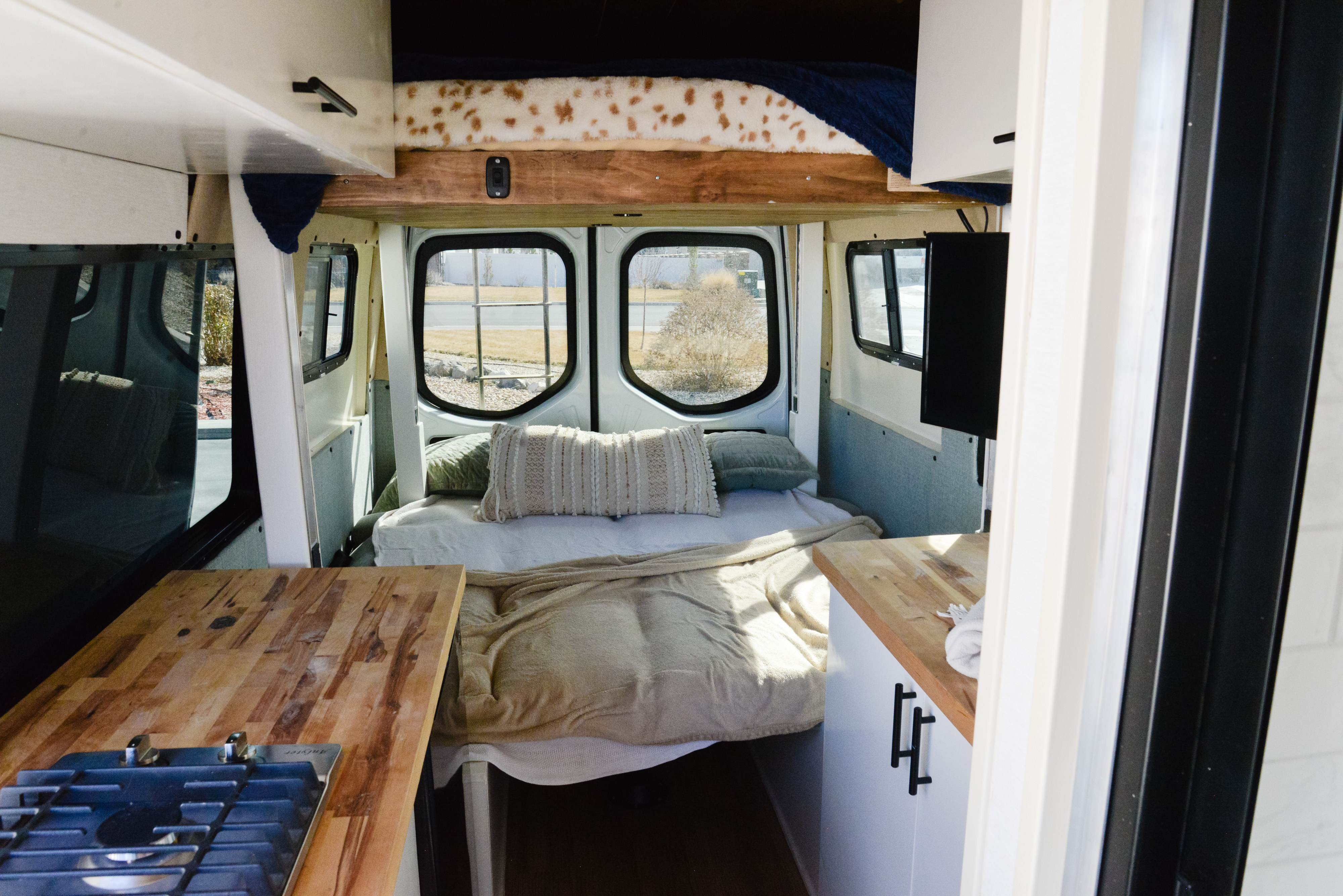 Inside a cozy rv rental camper van. Features a comfortable bed, kitchen counter with stove, and overhead storage.. Mercedes Sprinter 2022