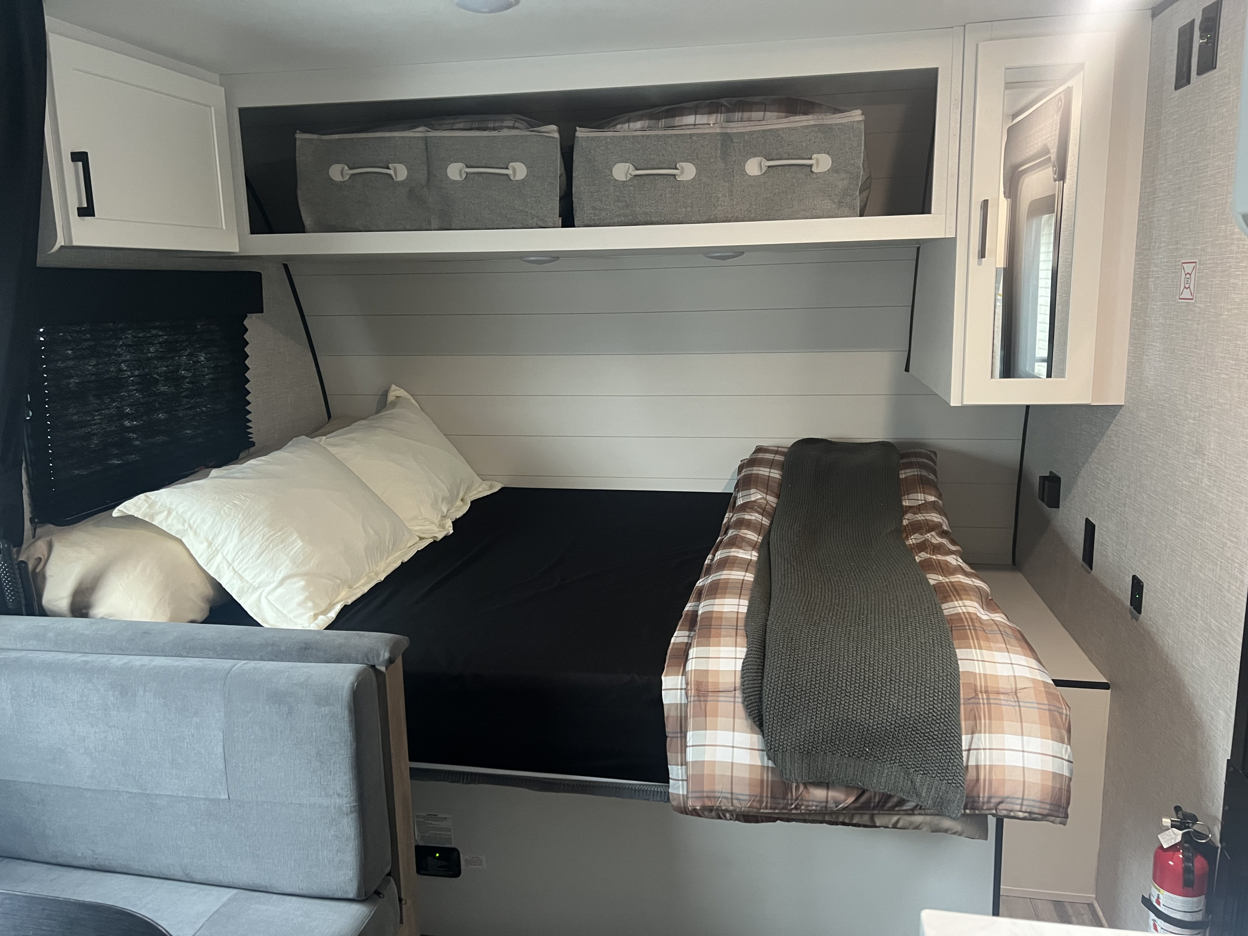 RV rental bedroom featuring a bed with white pillows, plaid comforter, overhead storage bins, and a grey sofa.. Jayco Jay Flight SLX Baja Edition 2025