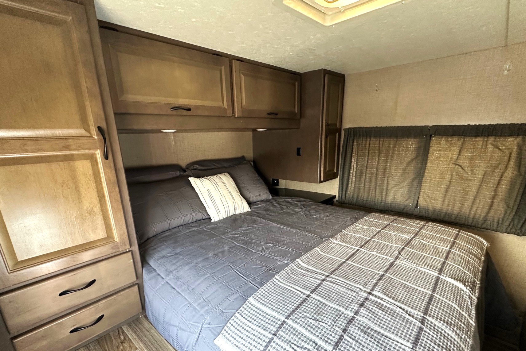 This RV rental bedroom features a cozy interior with a gray bed, overhead cabinets, wardrobe, and window curtains.. Thor Majestic 2019