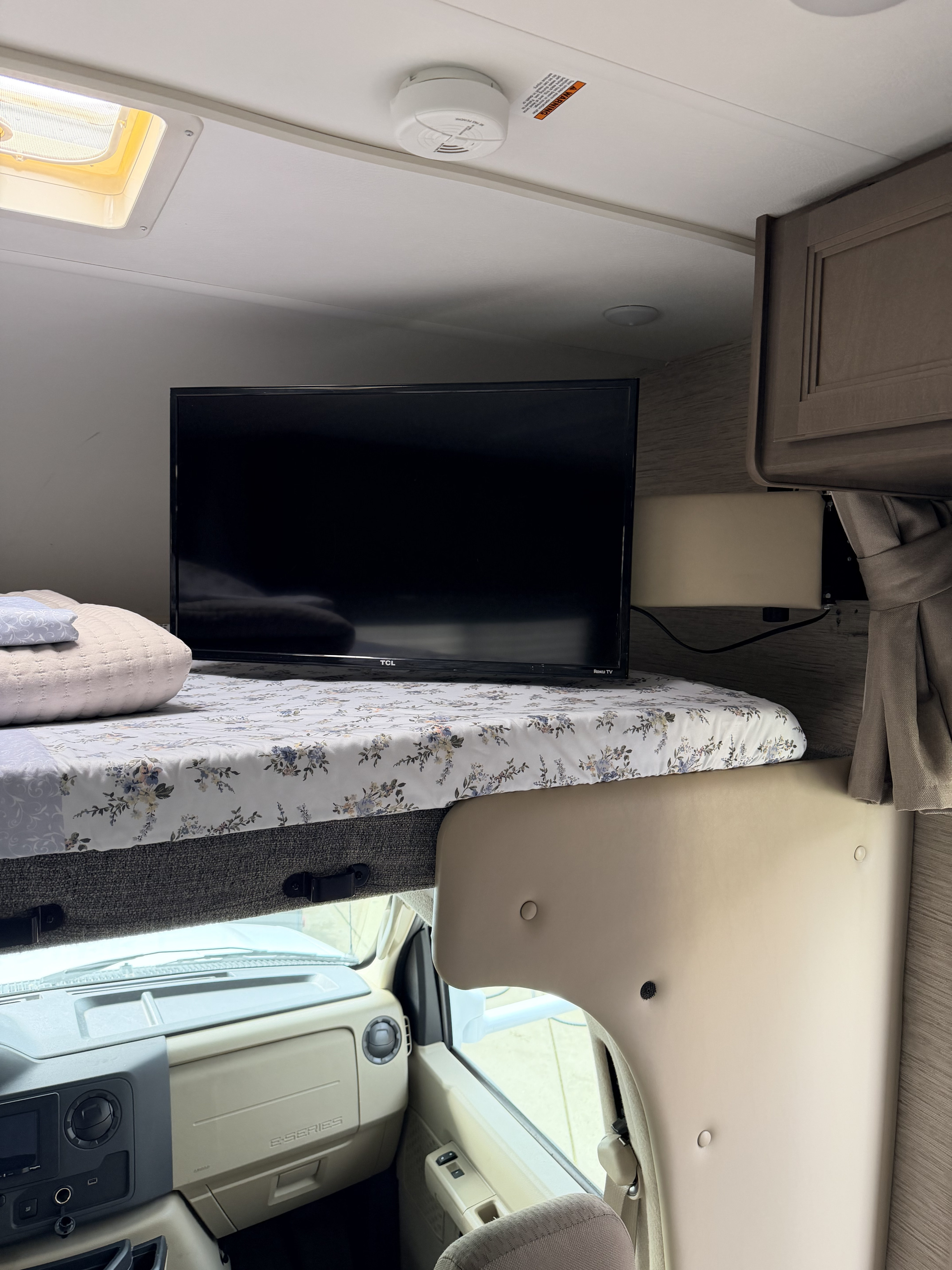 Inside an RV rental, showing the cab-over bunk with a TV, floral bedding, and the vehicle's dashboard below.. Thor Coleman 2023