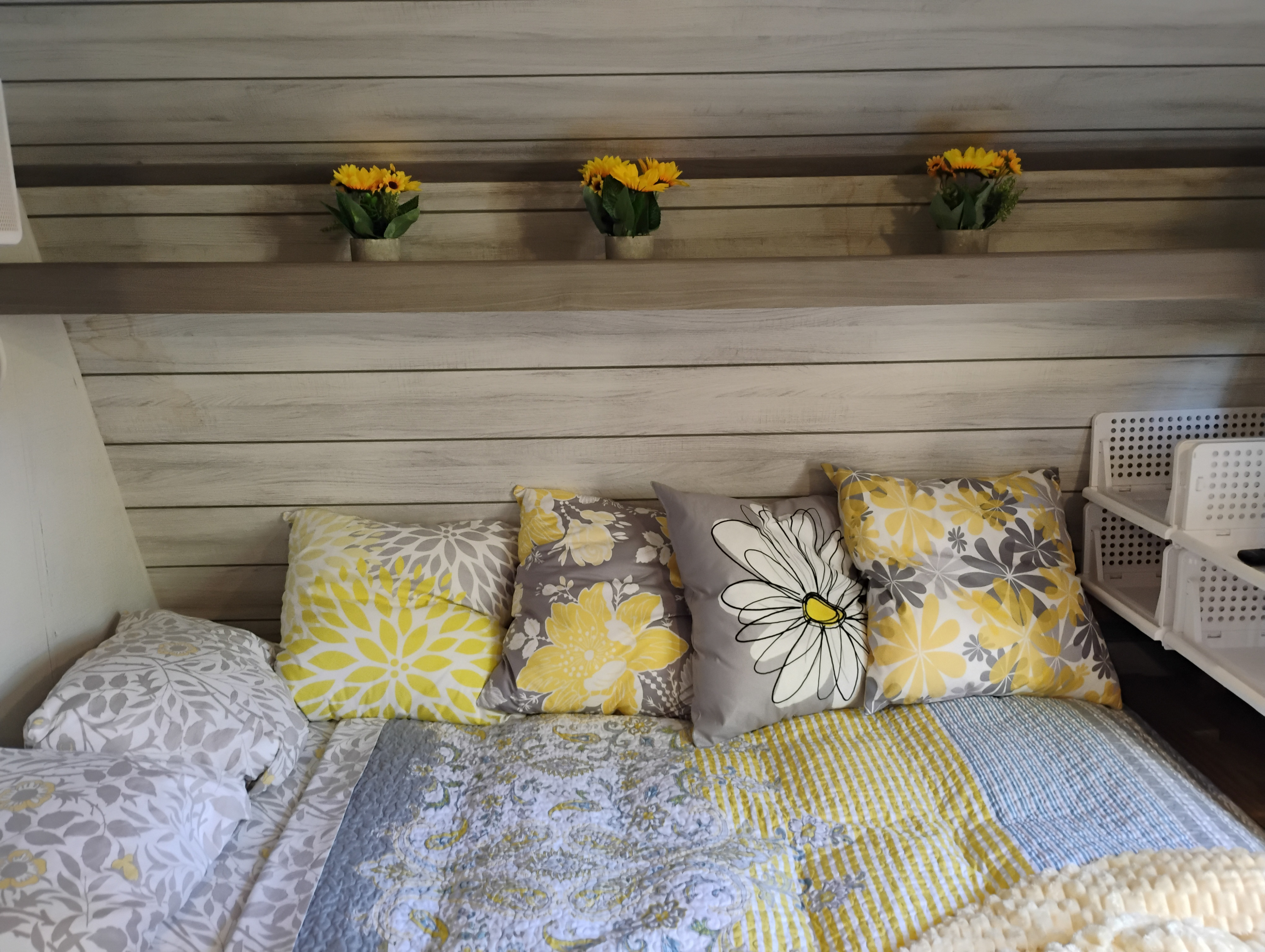 Cozy RV rental bedroom with yellow and grey floral pillows, quilted bedding, and decorative shelf with artificial sunflowers.. Dutchmen Coleman Lantern 2023