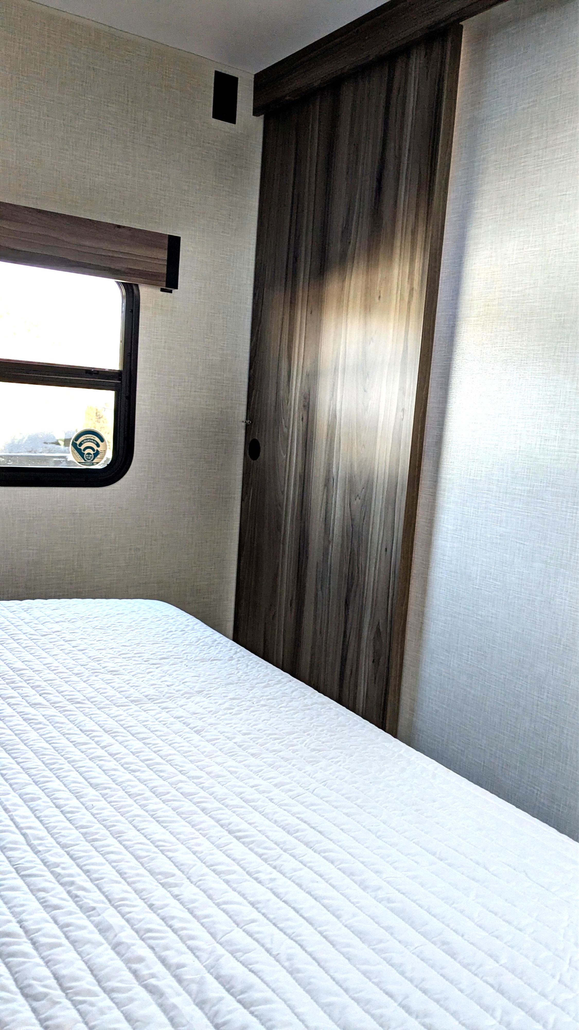 Cozy RV rental bedroom featuring a white quilted bed, window, and a modern wooden sliding door, offering a comfortable interior.. KZ Connect SE 2025