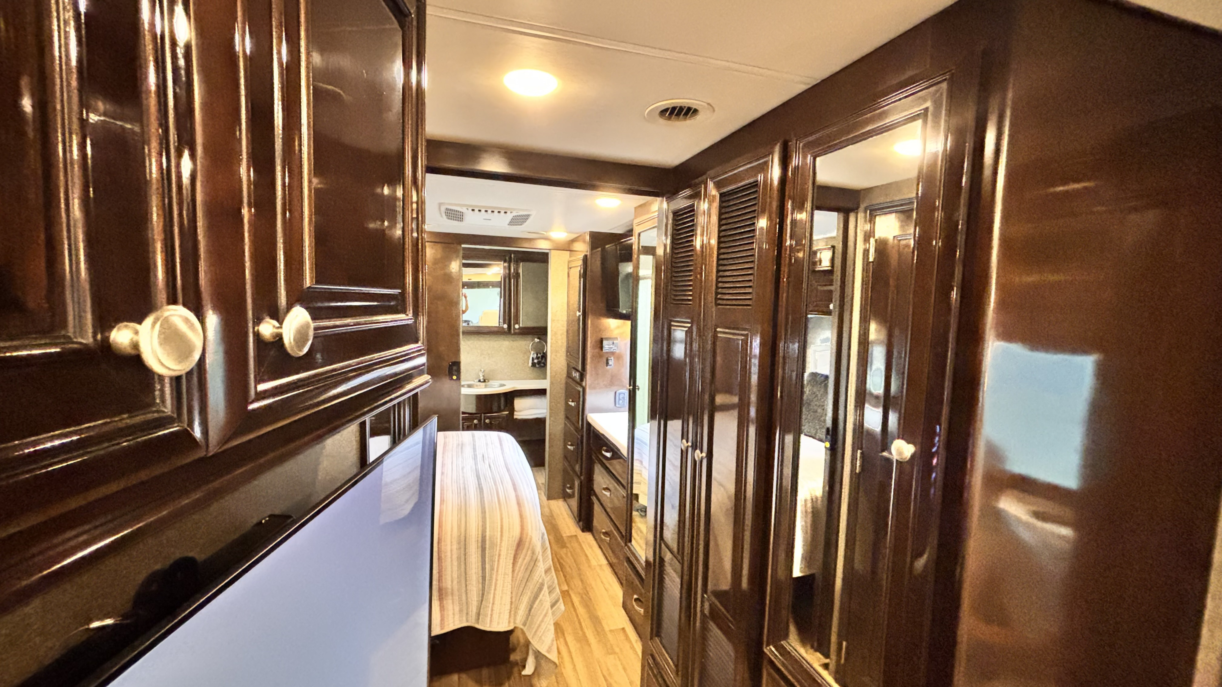 View of an rv rental's elegant dark wood interior hallway, leading past a bed and TV toward a bathroom area. Brightly lit.. Thor Miramar 2017