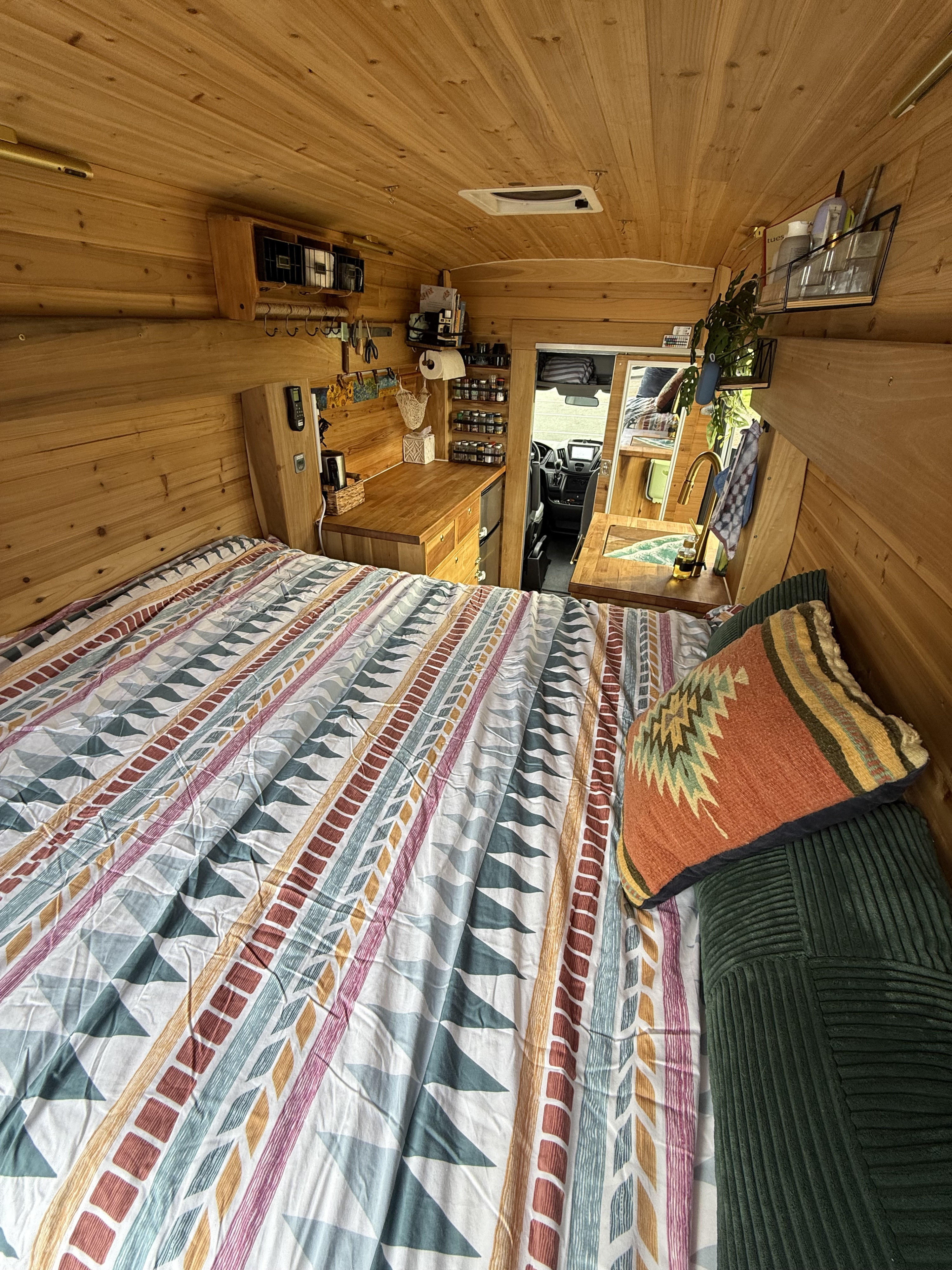 Cozy RV rental interior with a patterned bed, warm wood paneling, and a compact kitchen area with sink and storage.. Ford Transit 2019