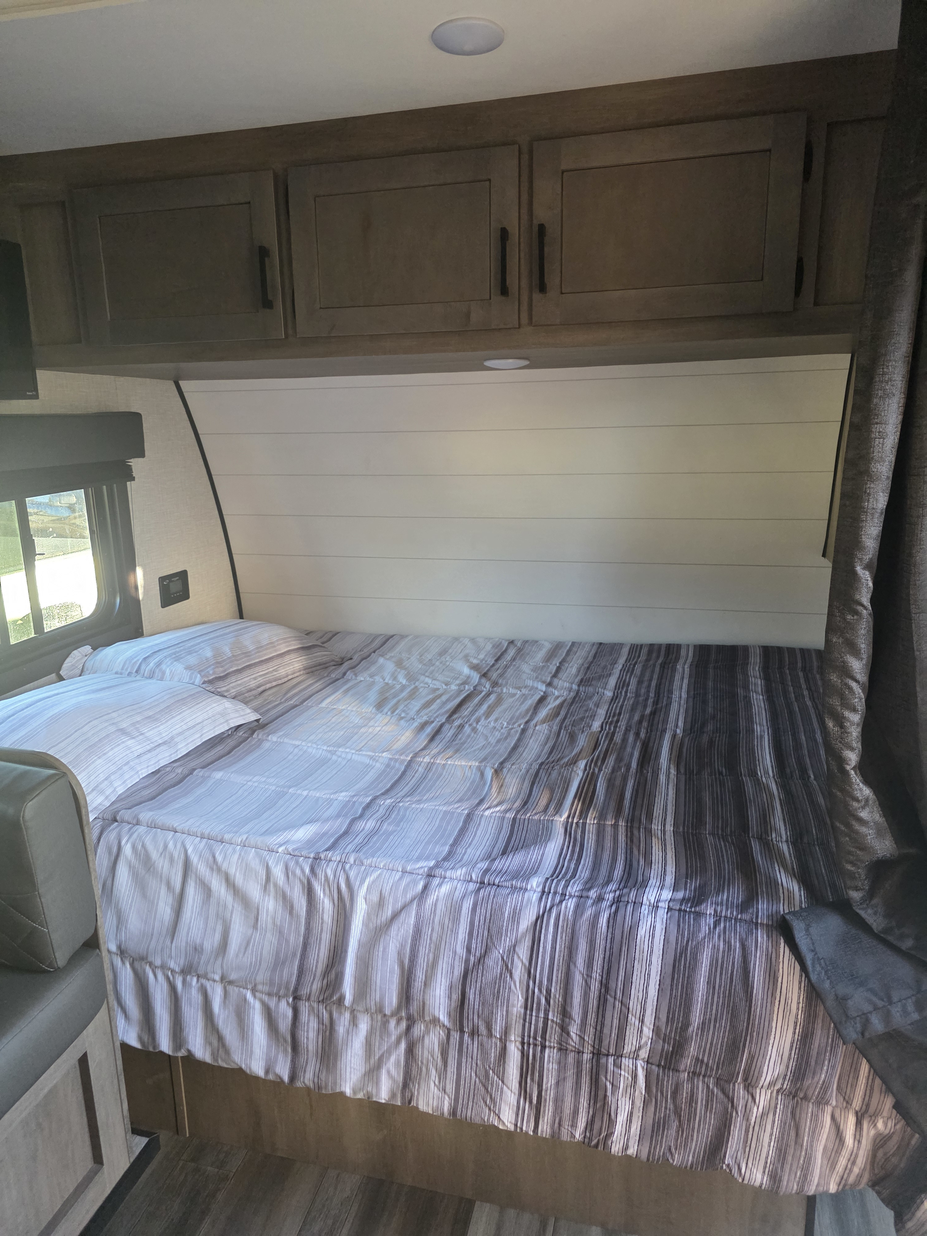 Cozy RV rental bedroom featuring a striped bed, shiplap headboard, overhead cabinets, and a window on the left.. Starcraft Autumn Ridge 2024