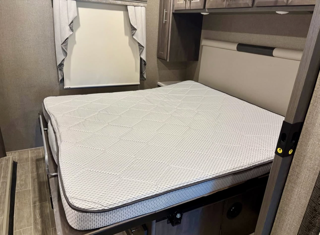 Cozy RV rental bedroom features a clean mattress, built-in headboard, and overhead cabinets, alongside a window with blinds.. Thor Miramar 2022