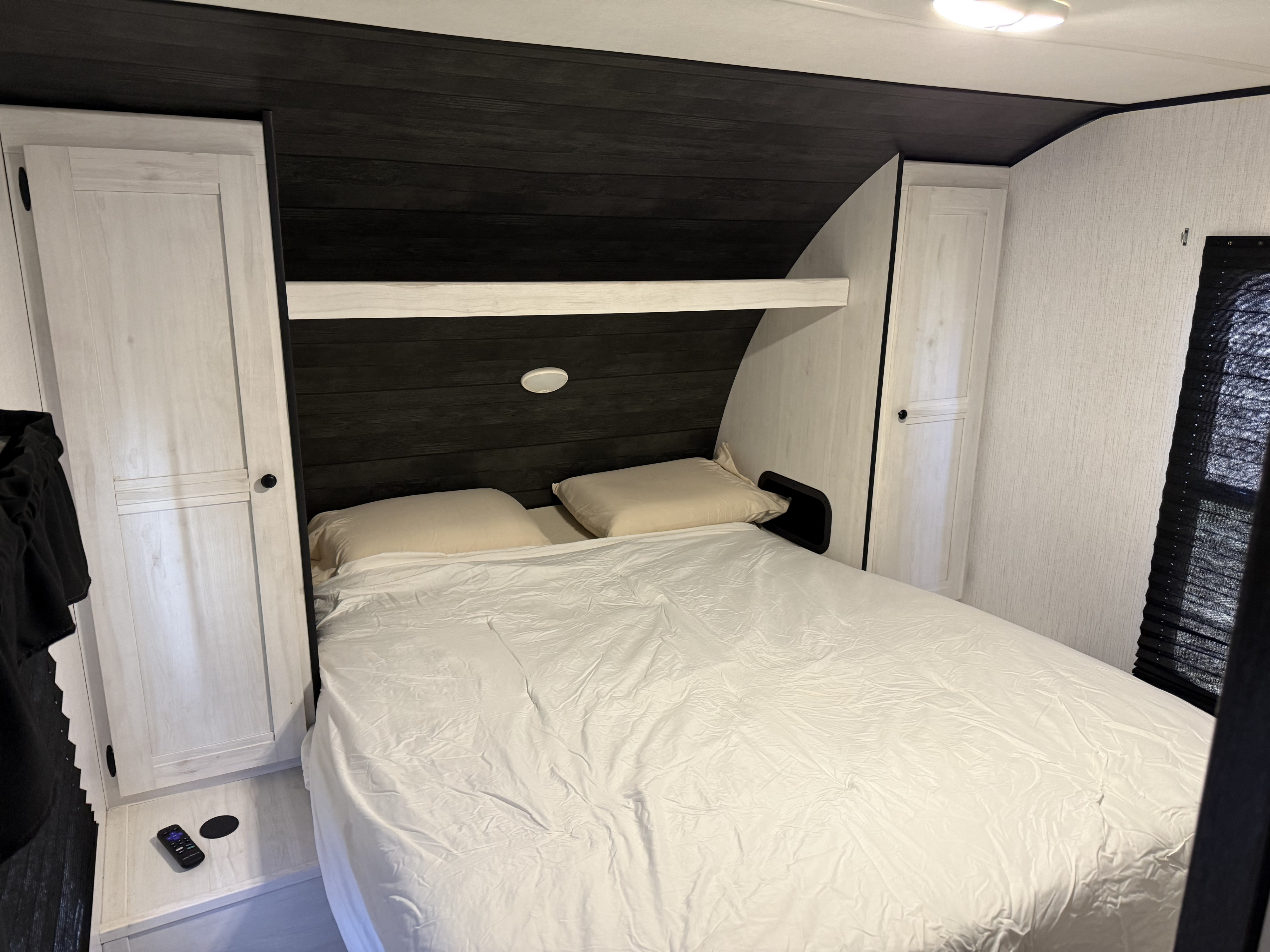 This rv rental interior features a compact bedroom with a bed, white sheets, dark wood paneling, light cabinets, and a window blind.. Heartland RVs Trail Runner 2022