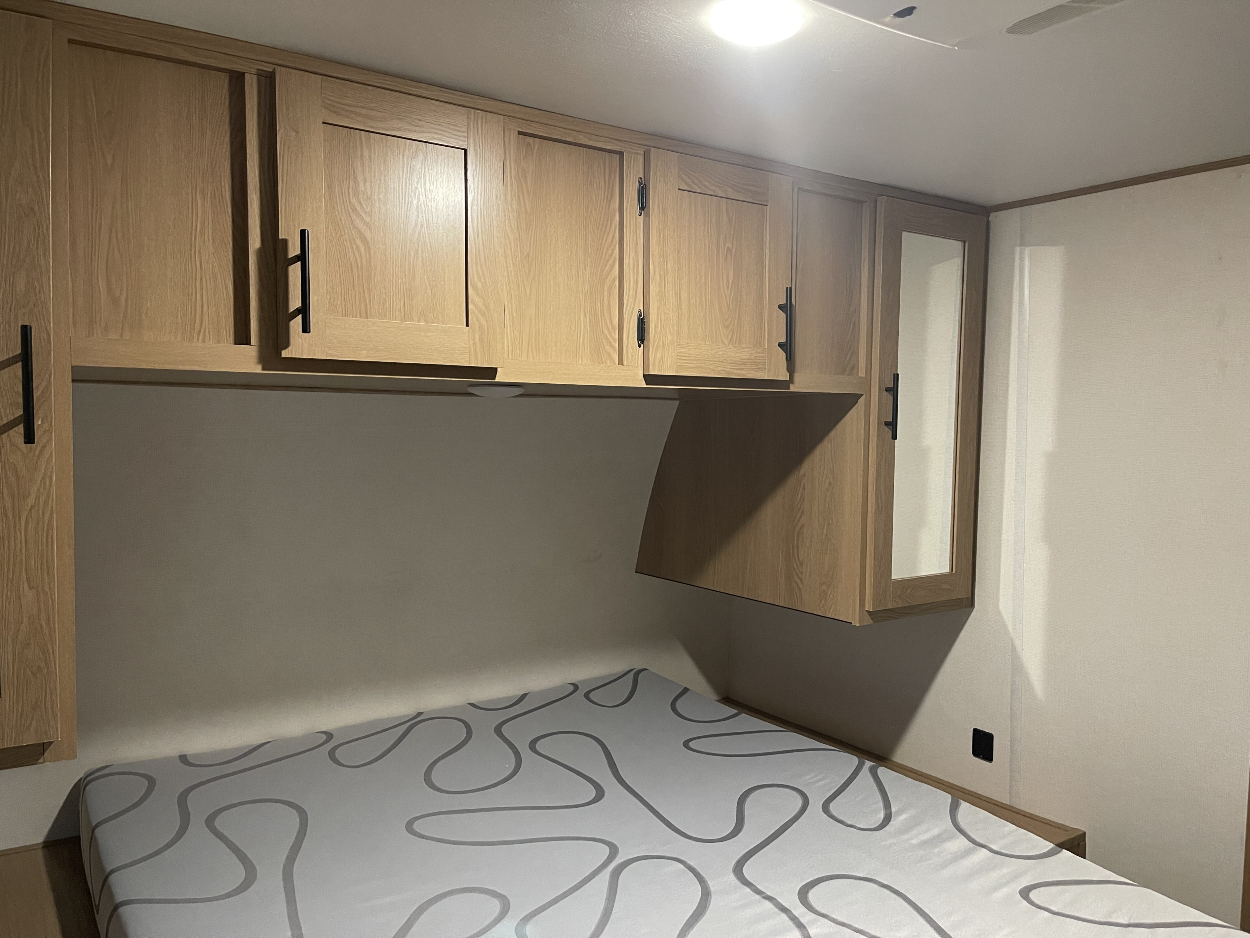 Inside an RV rental bedroom: light wood overhead cabinets, one with a mirror, above a bed with a grey patterned mattress.. Avenger Avenger Trailer 2022