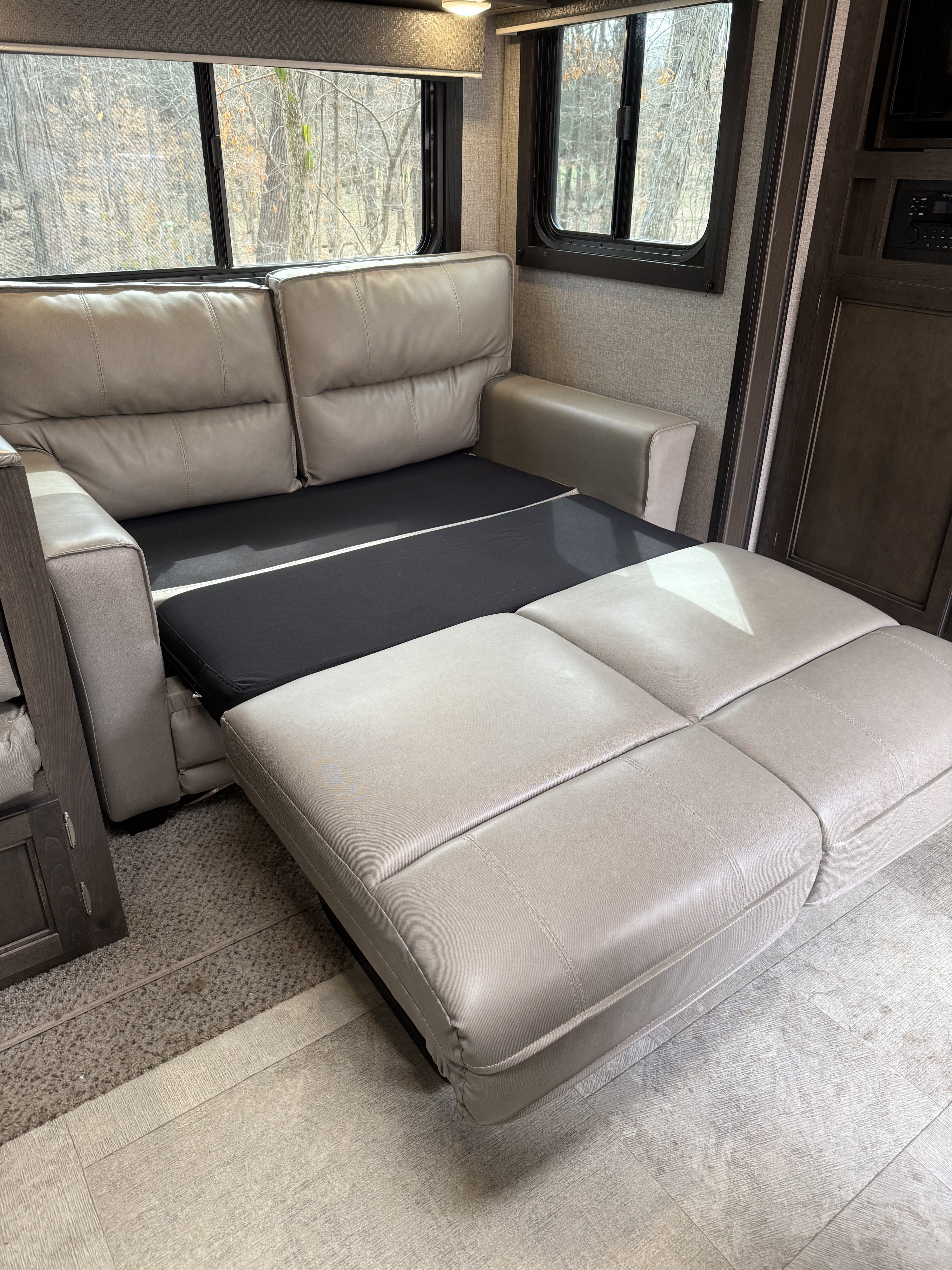 Inside an rv rental: a light gray sofa bed, partially extended for sleeping, by windows looking out to a wooded view.. Jayco Jay Flight 2020