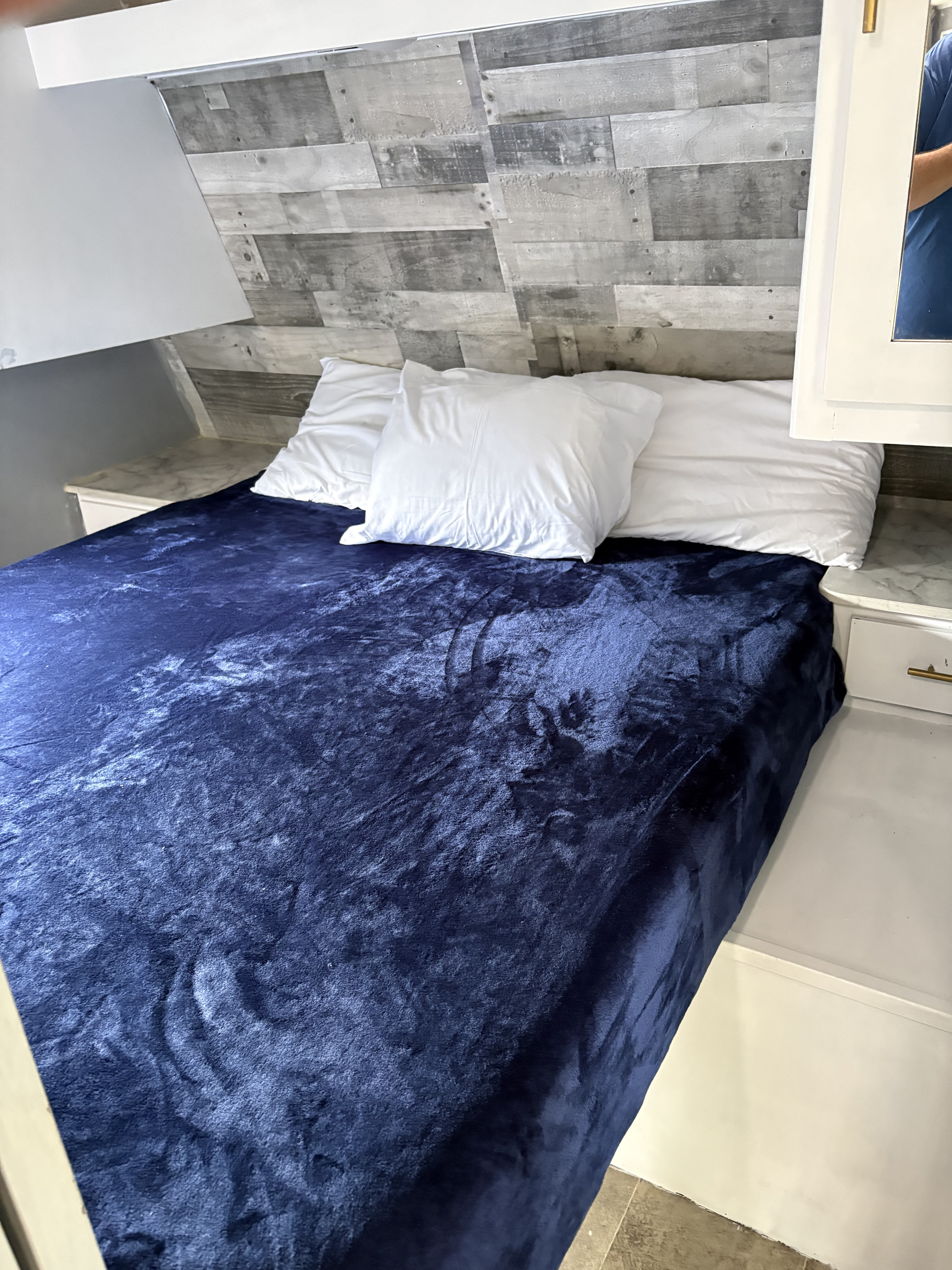 Inviting RV rental sleeping space features a deep blue blanket, white pillows, and a rustic wood accent wall. Compact comfort for travel.. Dutchmen Coleman Lantern 2021