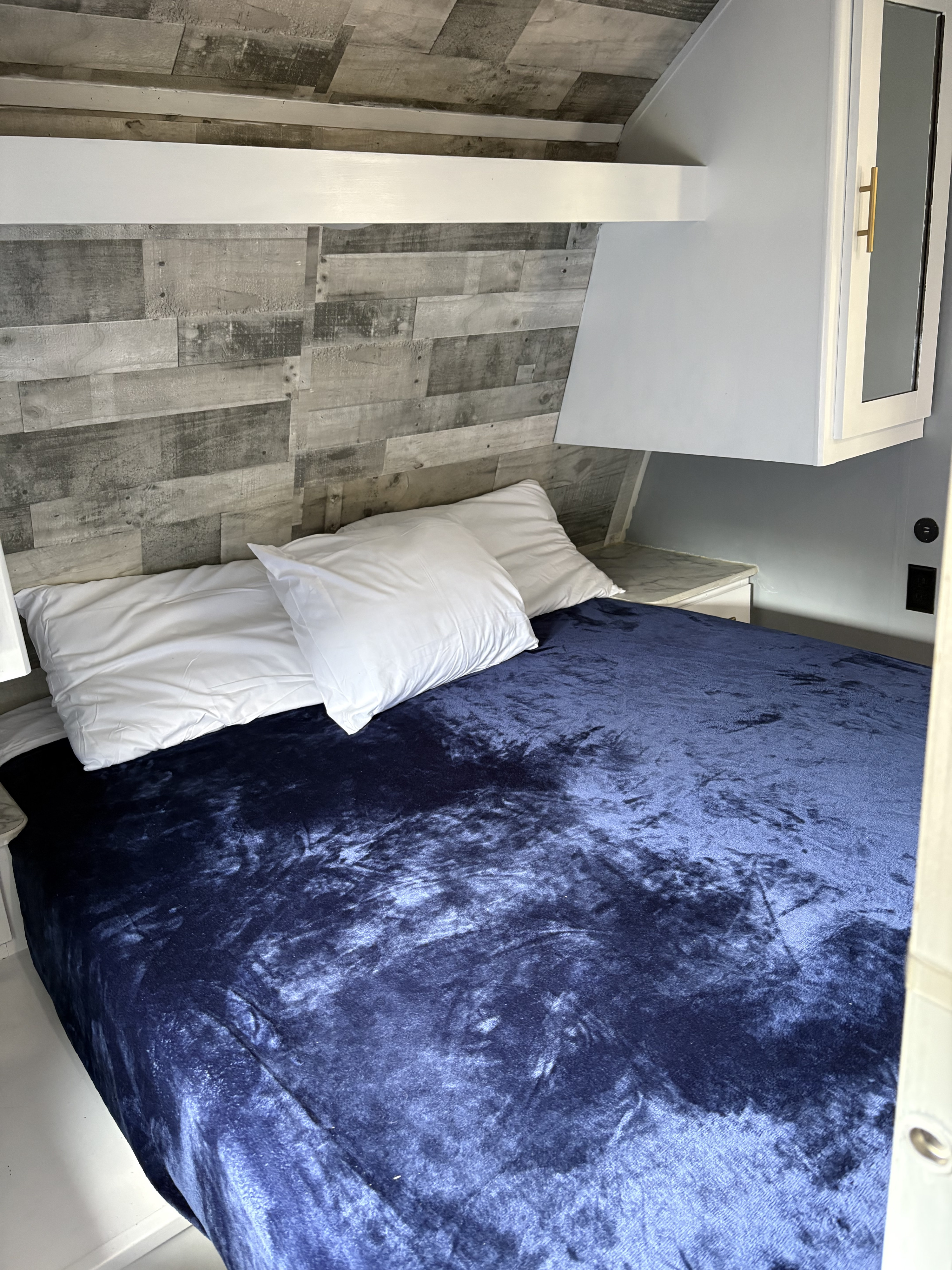 Cozy RV rental bedroom with a plush blue velvet blanket, white pillows, and rustic wood-look walls, ideal for comfortable travel.. Dutchmen Coleman Lantern 2021