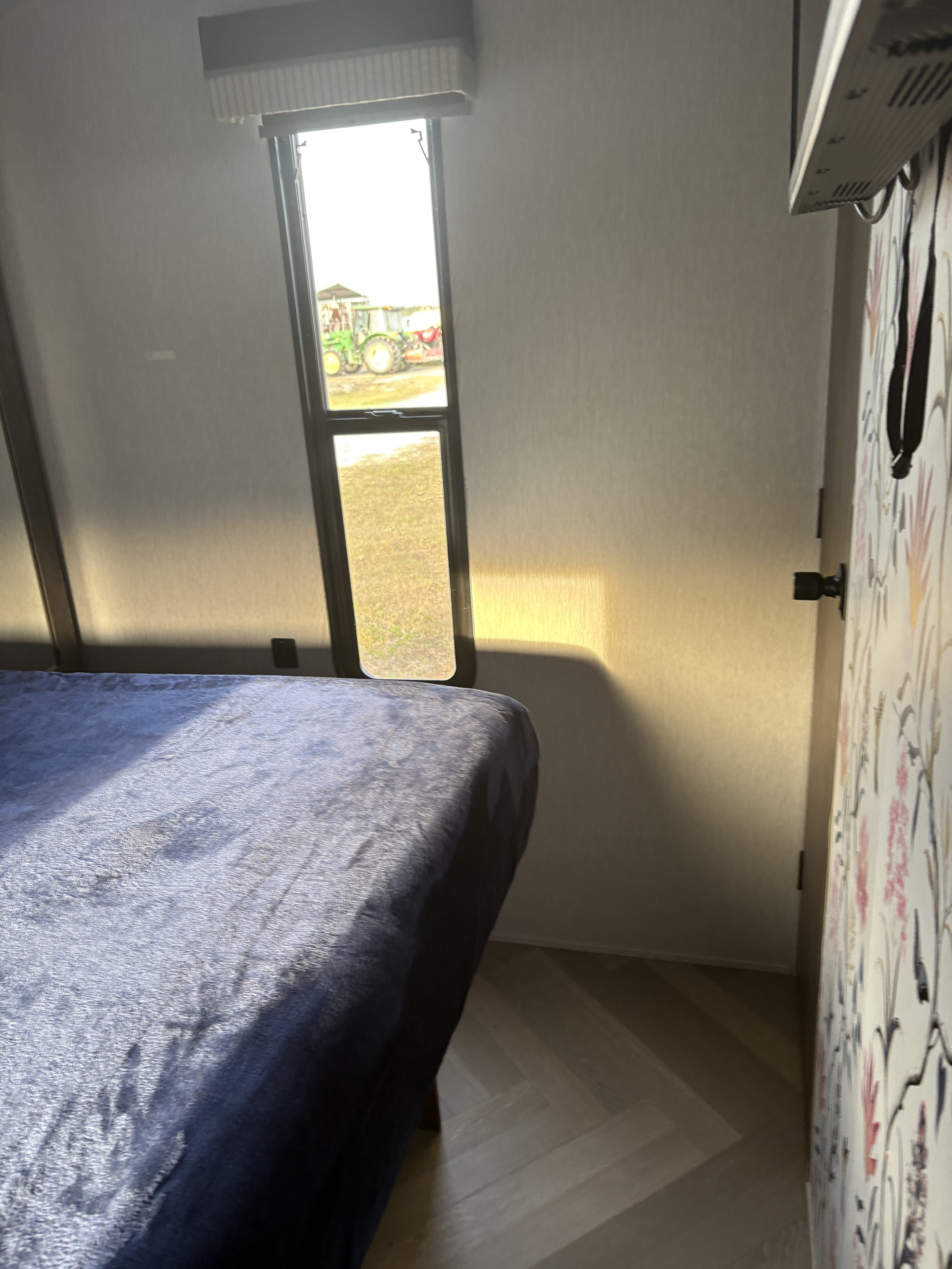 RV rental bedroom with a blue bed, sunlit window view of a green tractor, and patterned door.. Forest River Wildwood 2022
