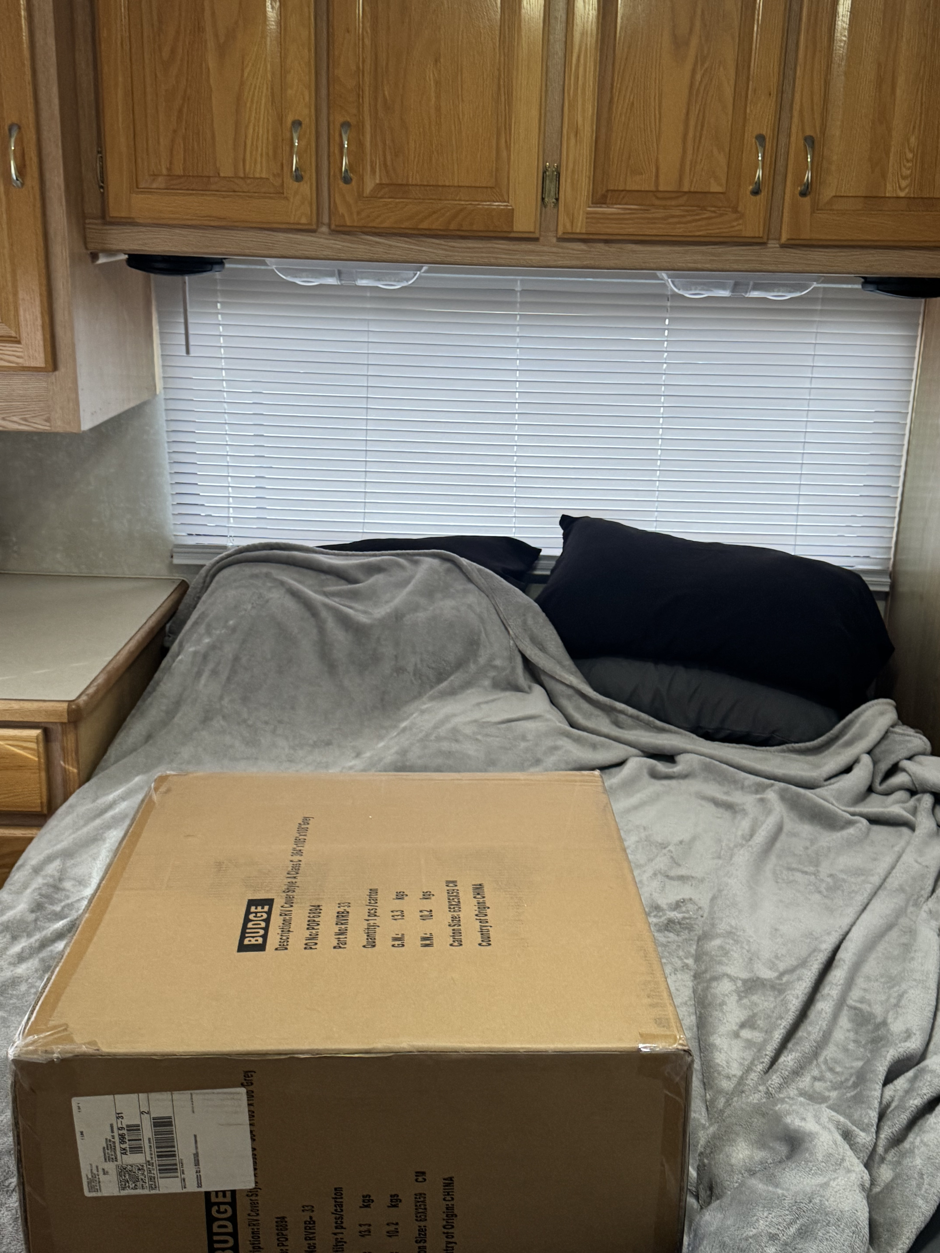 Inside an RV rental, a boxed Budge RV cover sits on a cozy bed with grey blankets and pillows, below wooden cabinets and blinds.. Forest River Sunseeker LE 2004