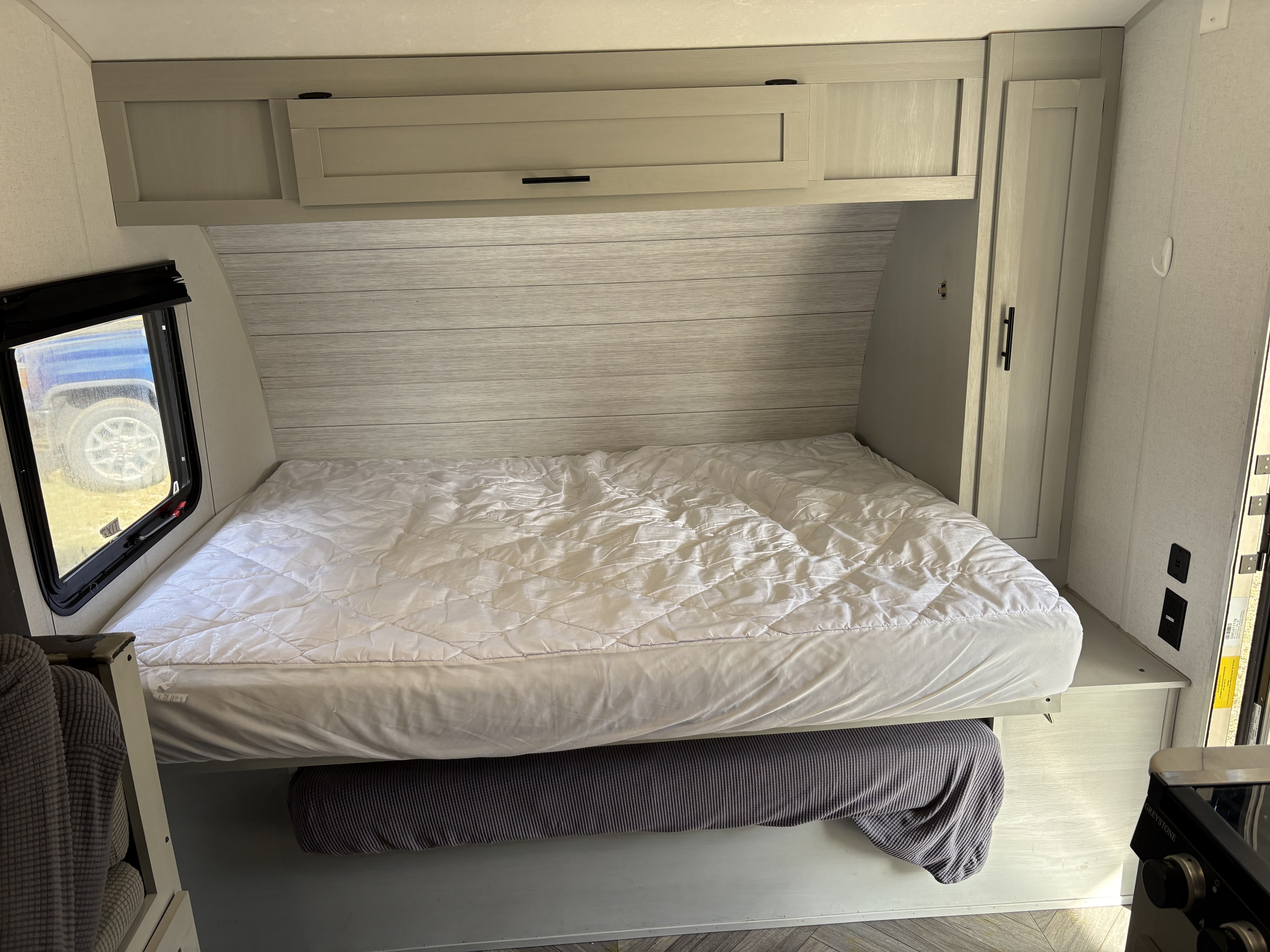 Clean RV rental bedroom with a white quilted bed, light wood paneling, overhead storage, and a side window view.. Forest River Salem FSX 2021