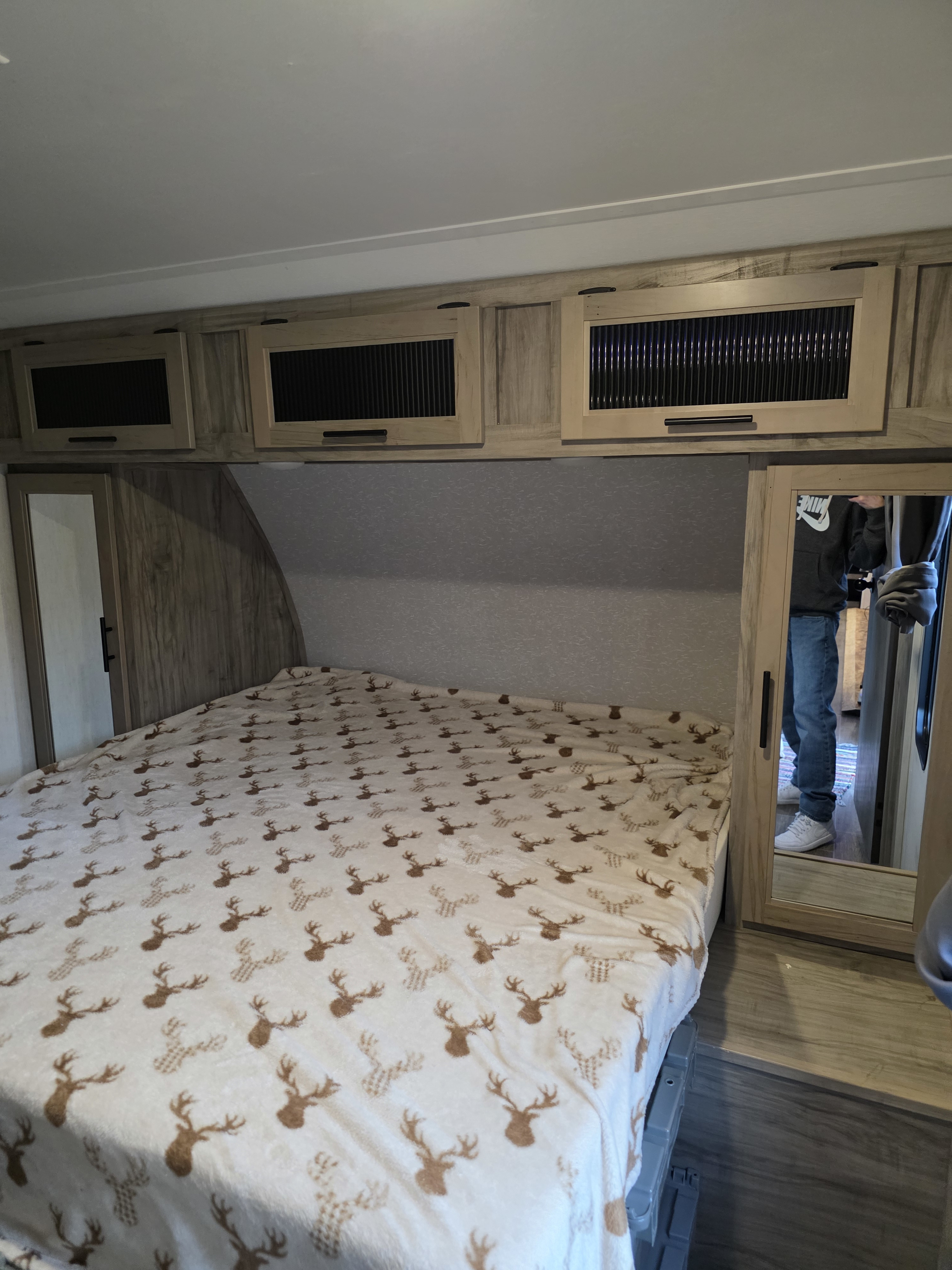 Cozy RV rental bedroom features a bed with a deer blanket, overhead cabinets, and a mirrored closet reflecting a person.. Forest River Cherokee Alpha Wolf 2022