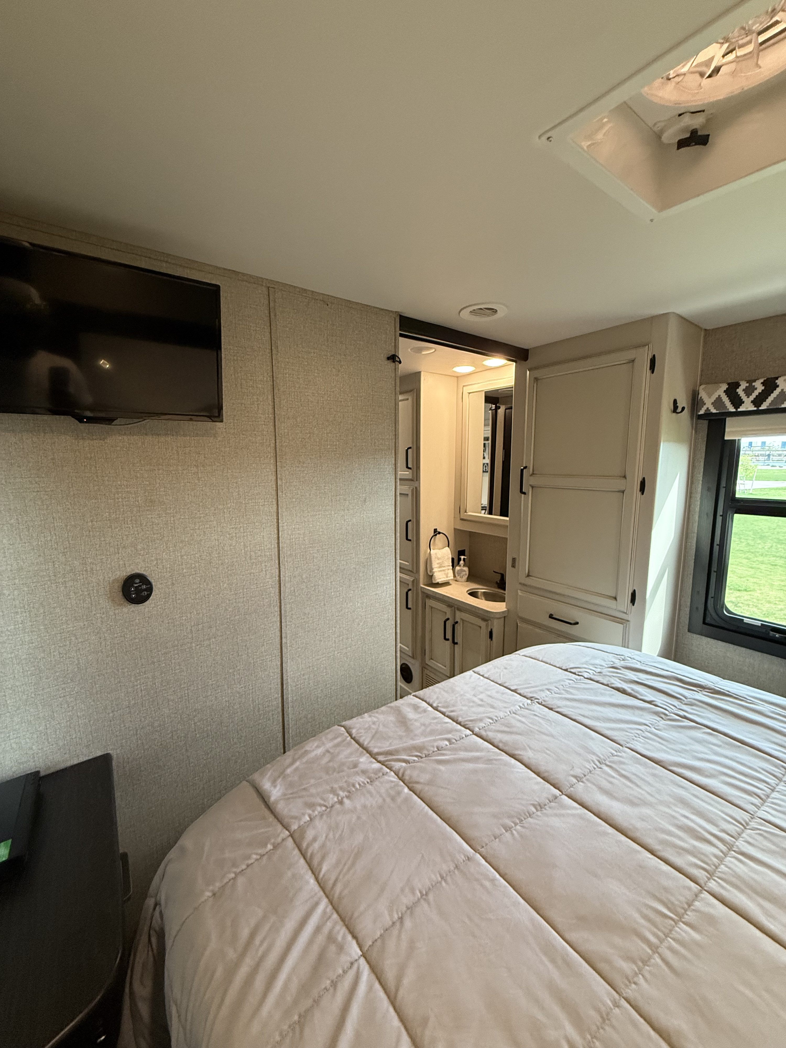 This RV rental bedroom features a comfy bed, wall-mounted TV, and a compact vanity/bathroom, perfect for travel.. Jayco Greyhawk 2021
