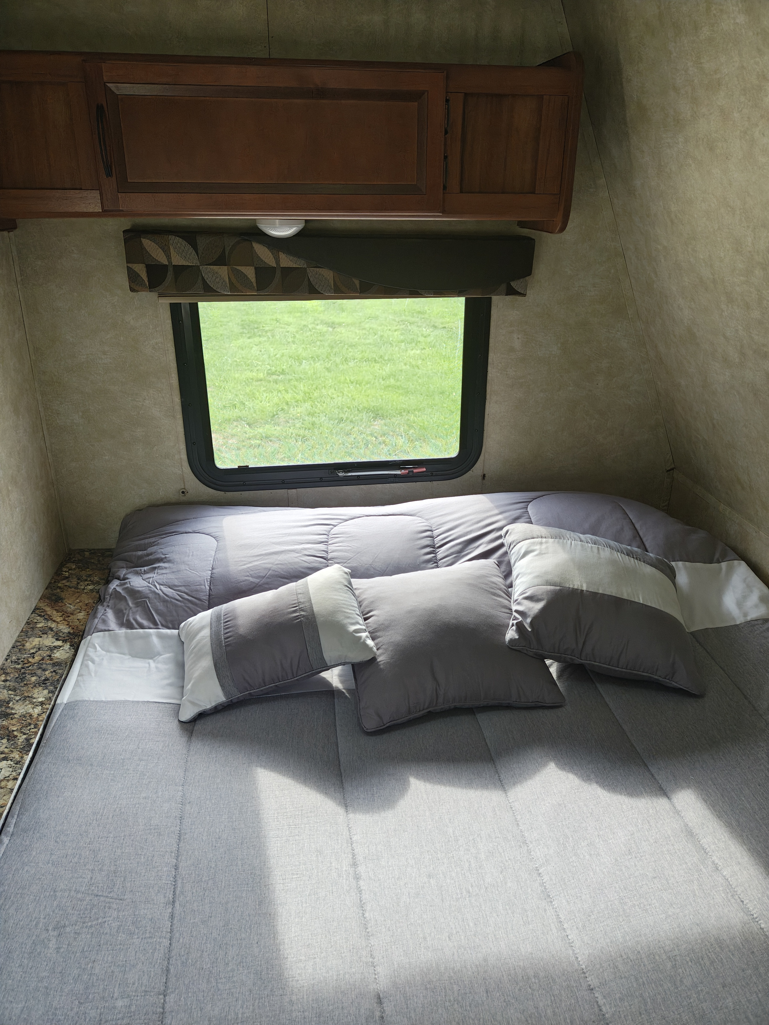 Cozy RV rental bedroom with a gray bed, pillows, overhead cabinets, and a window offering a lush green view outside.. Gulf Stream Track & Trail 2018