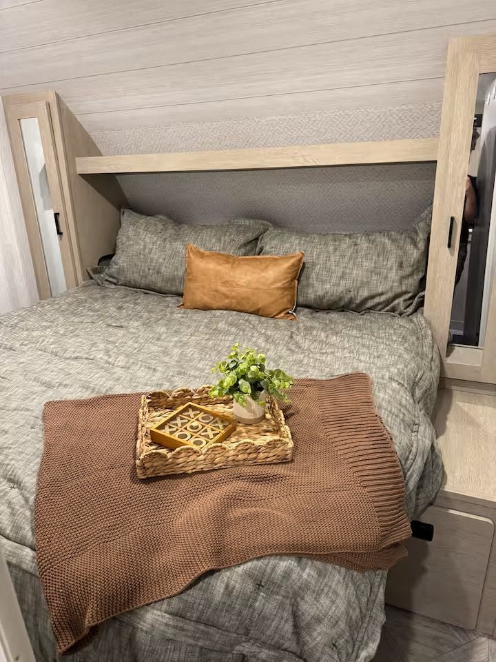 Cozy RV rental bedroom featuring gray bedding, a brown throw blanket with a tic-tac-toe game & plant, and light wood cabinetry.. Forest River Wildwood X-Lite 2023