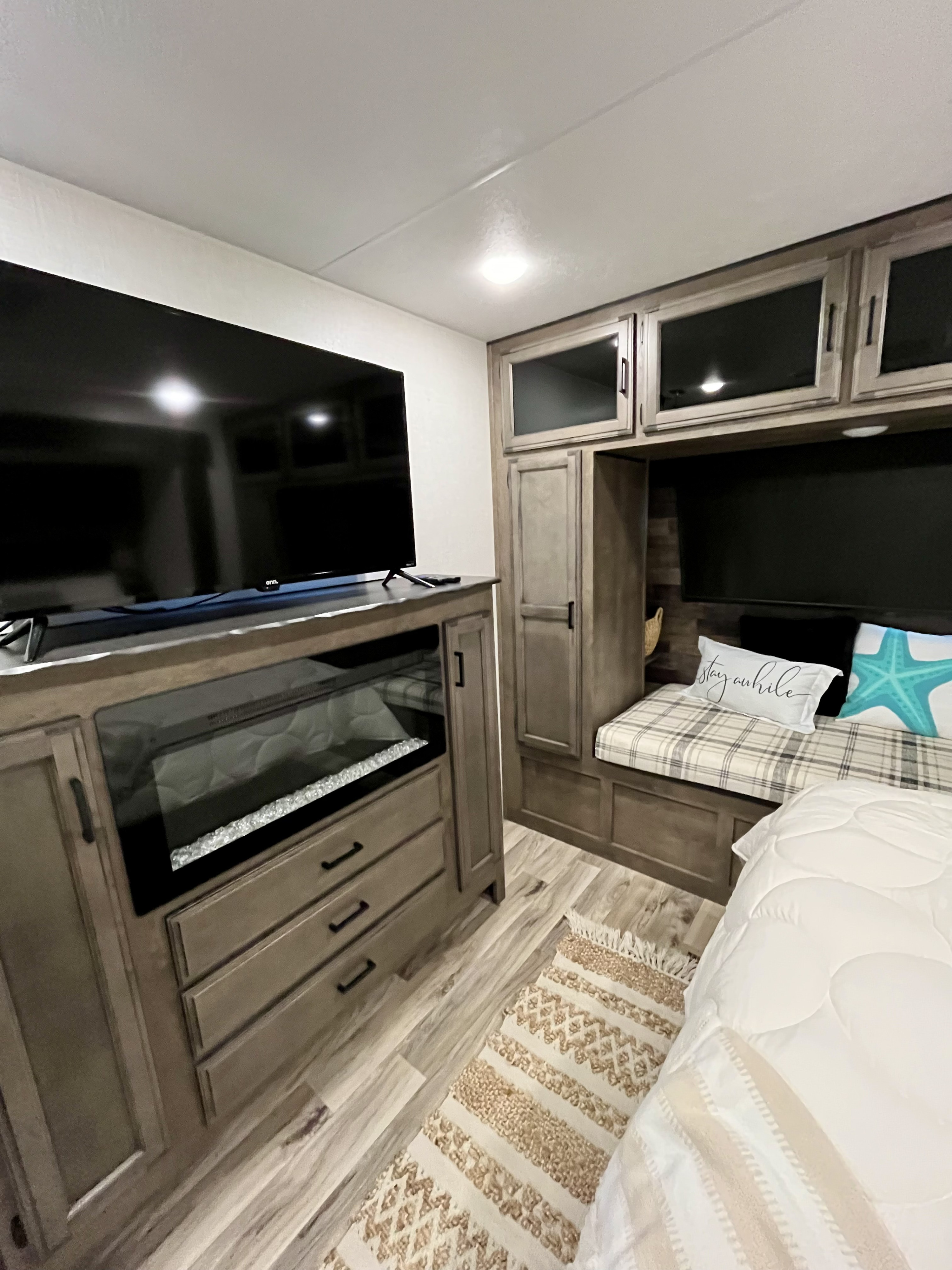 Inside a modern RV rental: a cozy bedroom with a large TV, electric fireplace, wood-look cabinets, padded bench, bed with white comforter.. Sporttrek Touring Edition 2024